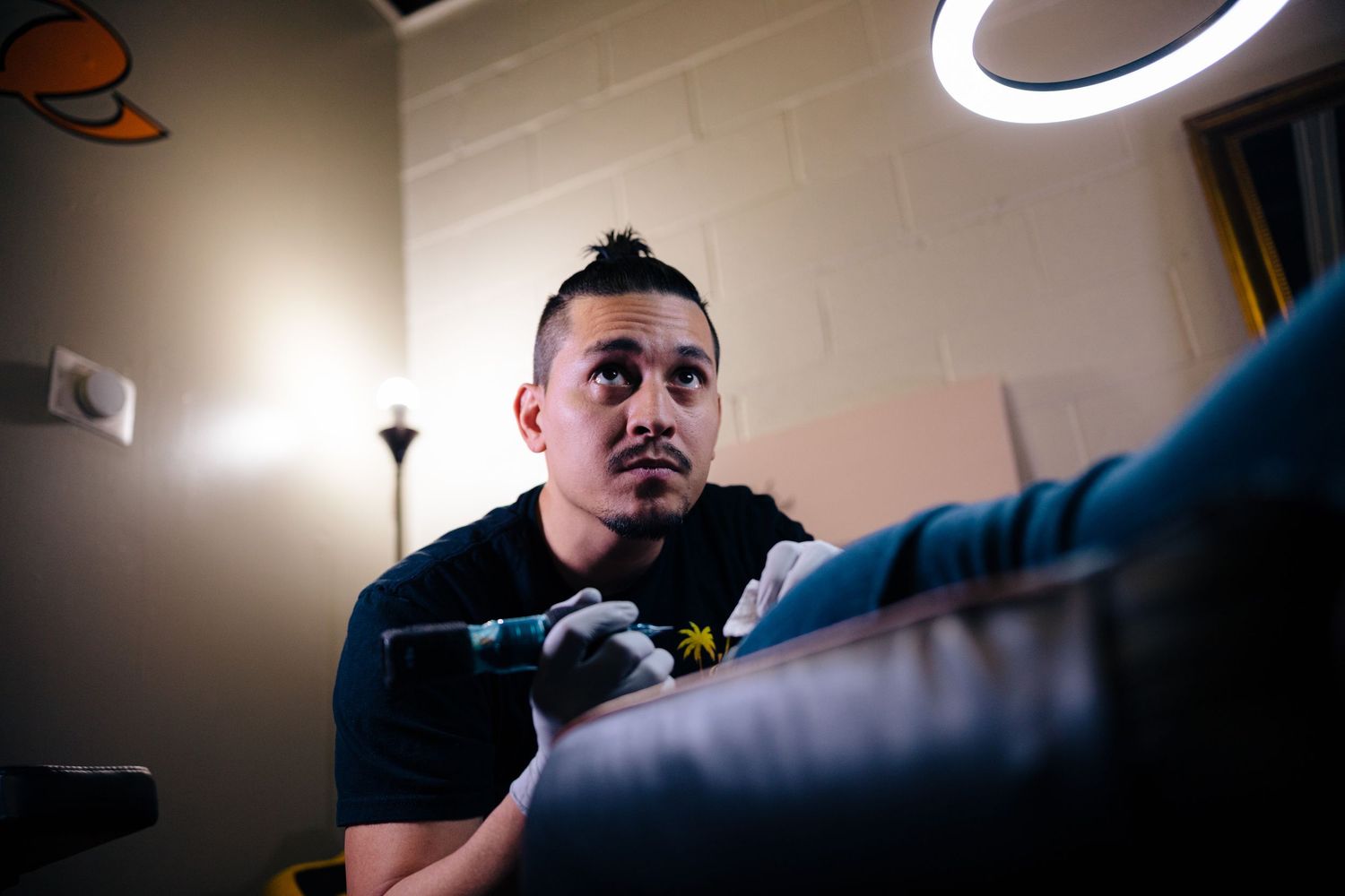 A focused tattoo artist applies ink to a client in a dimly lit studio, illuminated by a circular ring light overhead.
