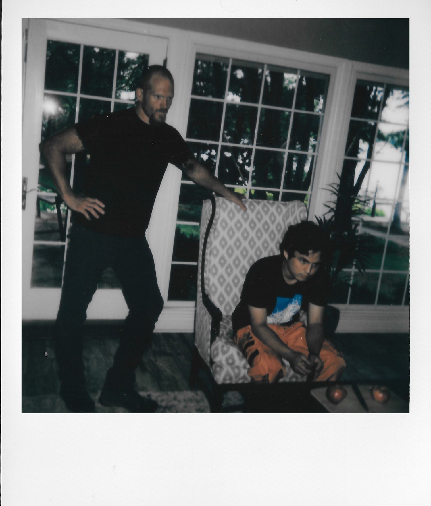 A bald man in black stands beside a boy sitting in a patterned armchair near large windows indoors.