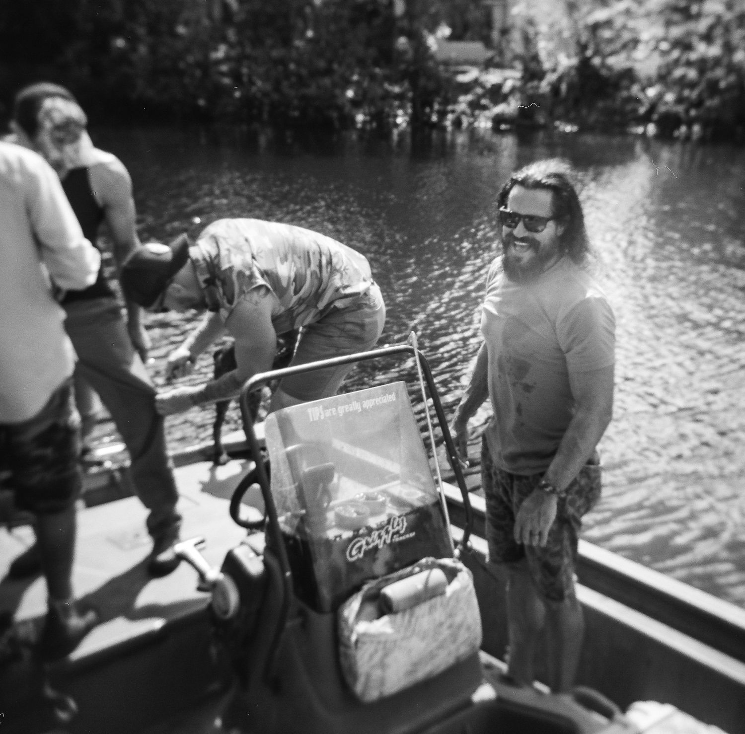 Black and white photo of people on a boat near a river, with a man in sunglasses smiling beside a stroller.
