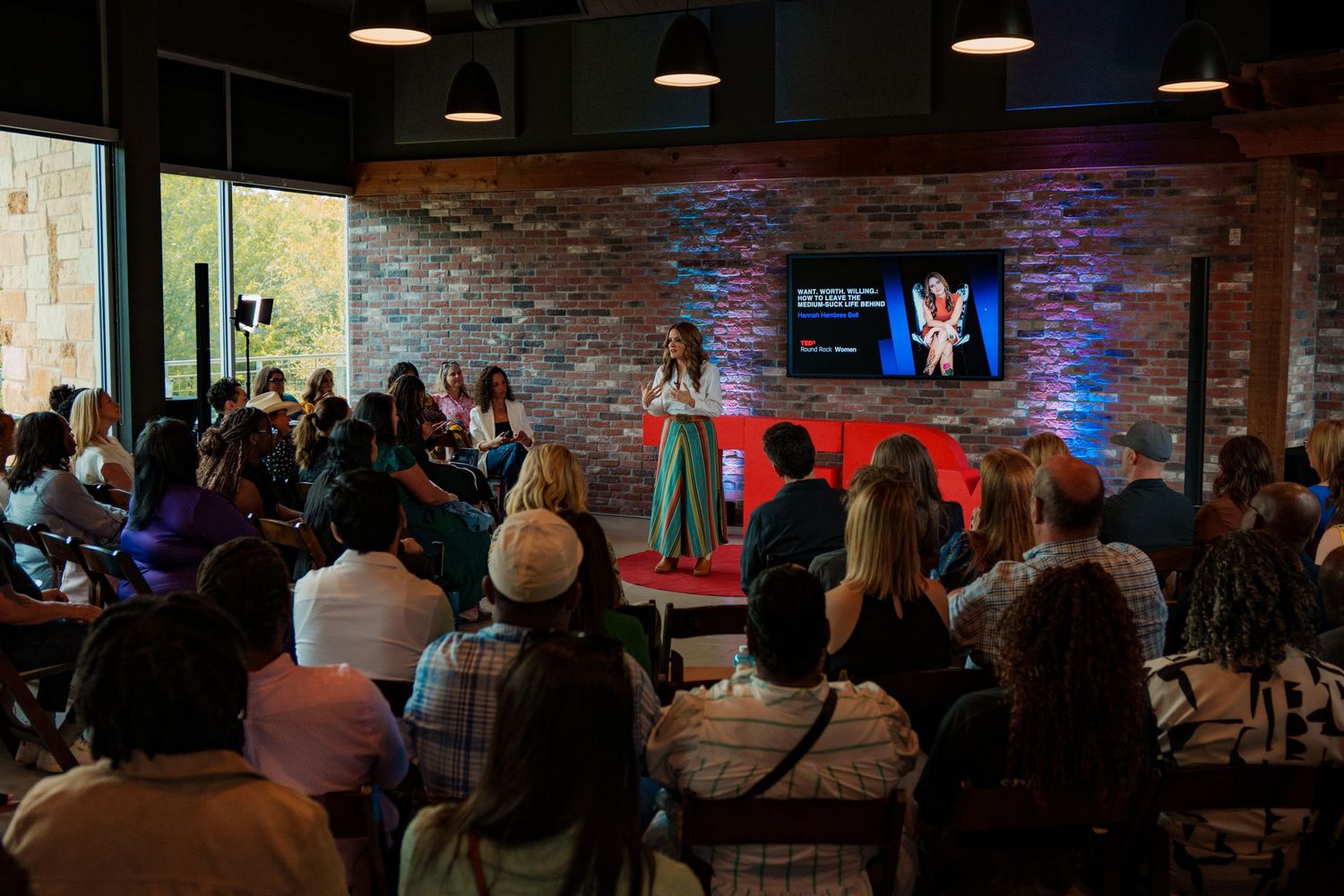 Hannah Hembree Bell presents at a TEDx event in a brick-walled venue with an engaged seated audience. Round Rock Texas.