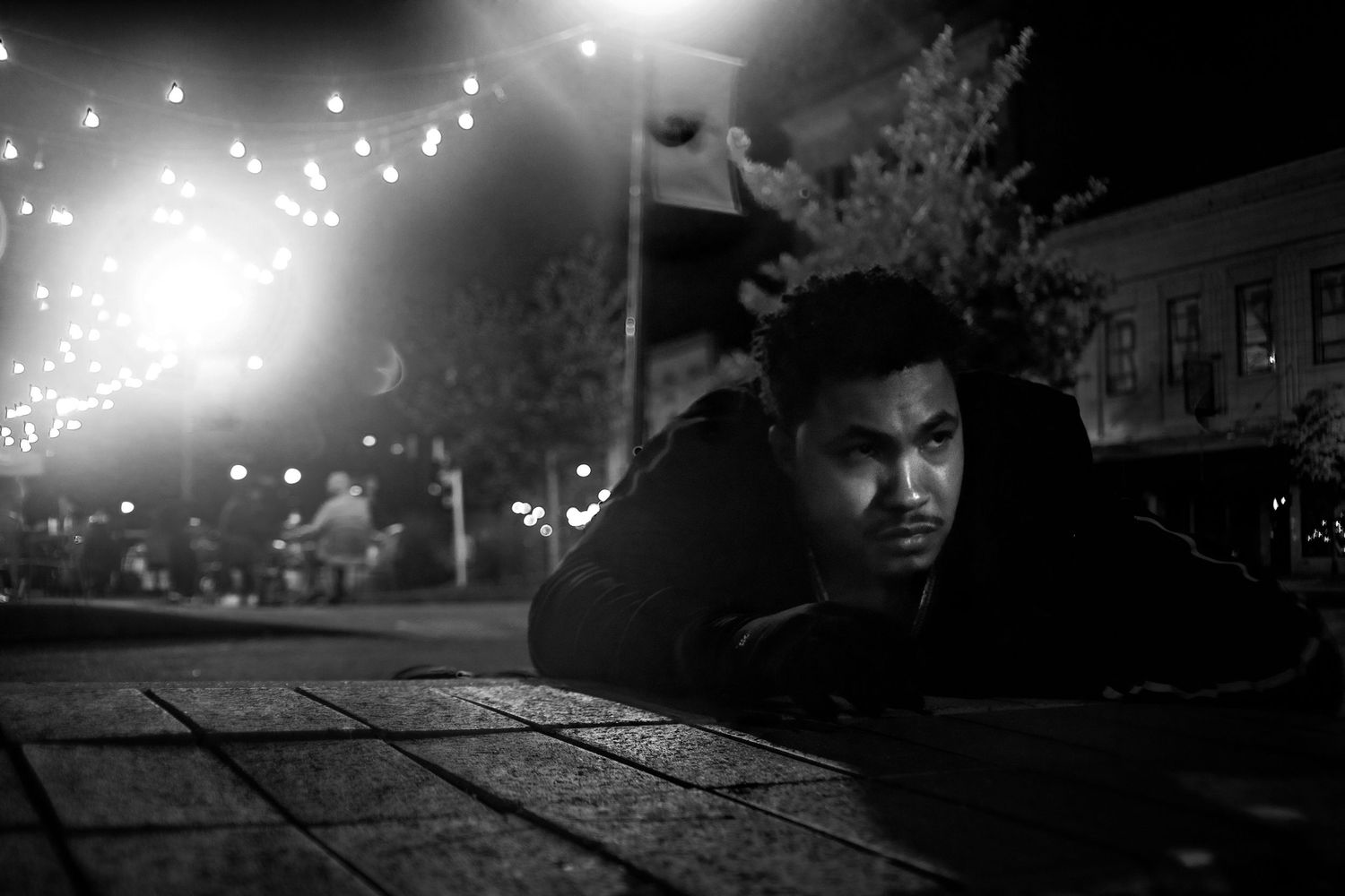 Black and white night photo of a young man lying on a sidewalk, looking up with city lights in background.