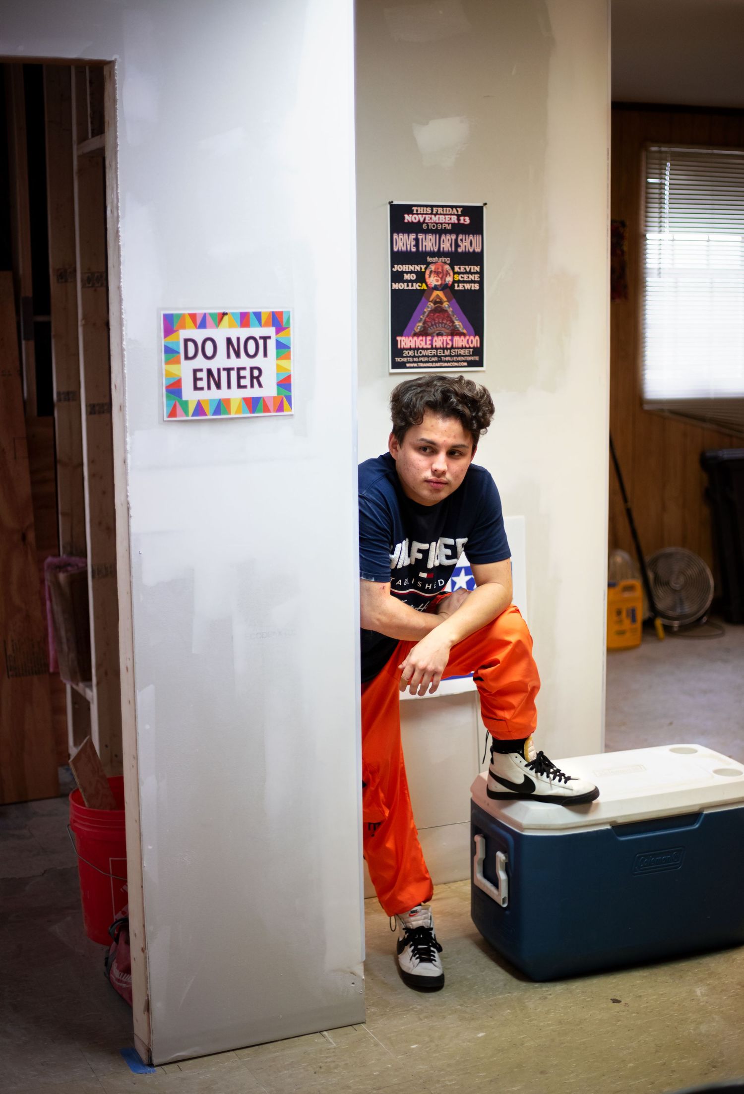 Teen boy in orange pants sits on cooler near a Do Not Enter sign in a garage with movie posters.