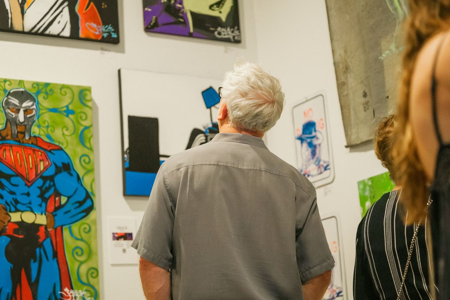 Gray-haired man viewing colorful artwork including a Superman painting at an indoor art gallery exhibition.