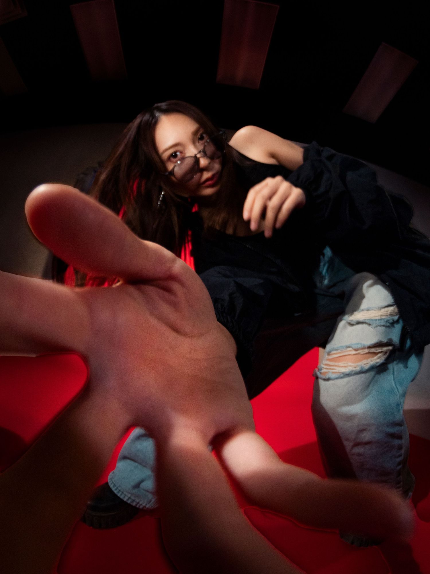 Woman in ripped jeans and glasses reaches toward camera in dramatic low-angle red-lit studio shot.