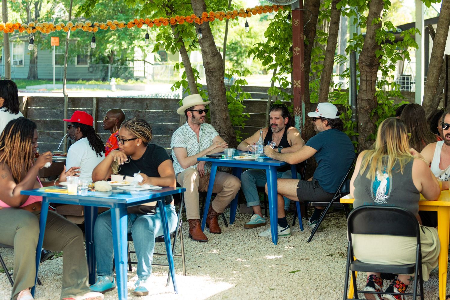 People enjoying outdoor dining under trees at blue tables during a sunny daytime gathering.