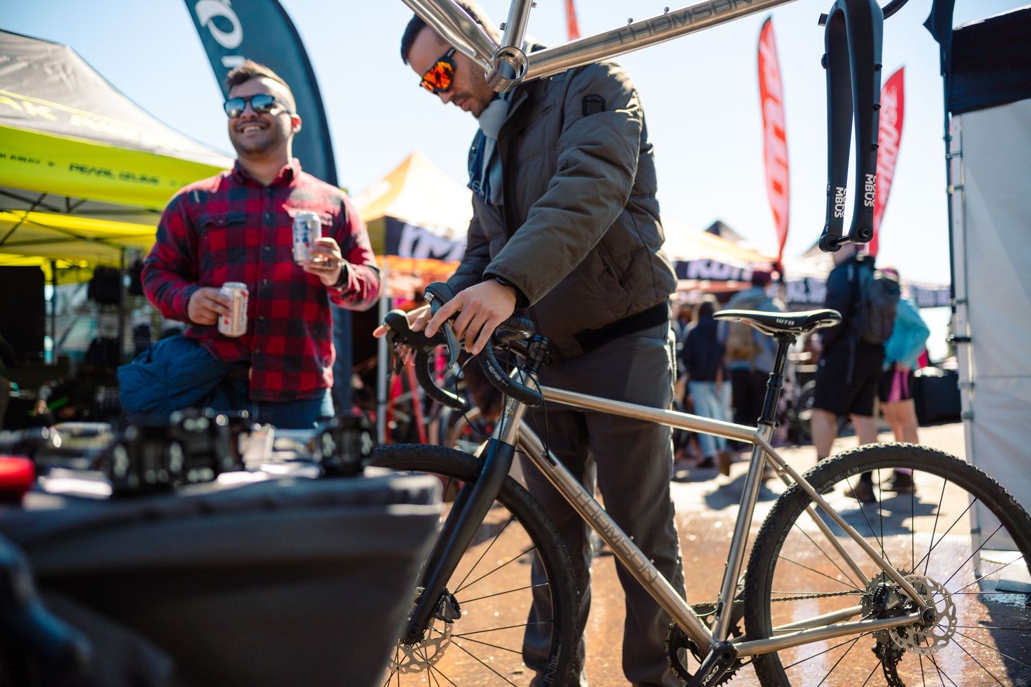 Man inspecting a titanium gravel bike at an outdoor cycling trade show with colorful vendor tents behind.