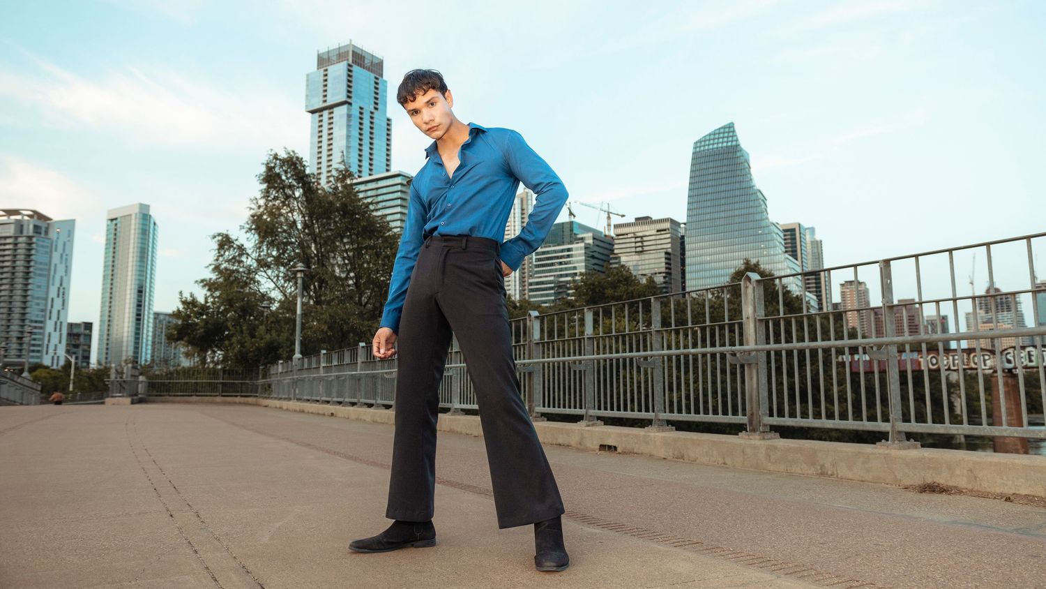 Man in blue shirt and black flared pants poses by city skyline.