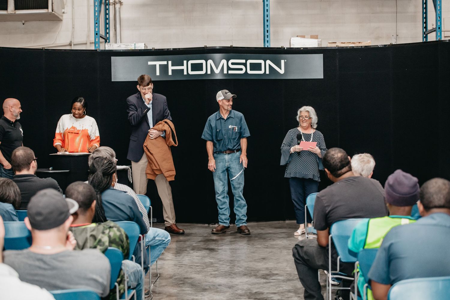 Thomson company employees gathered for a presentation ceremony in an industrial warehouse setting.