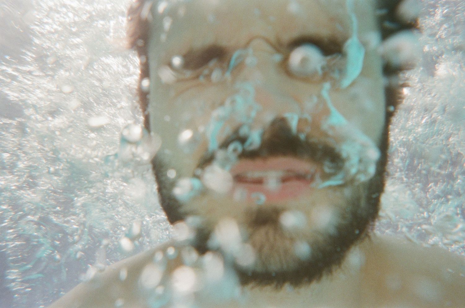Close-up underwater portrait of a bearded man submerged in water with bubbles surrounding his face.