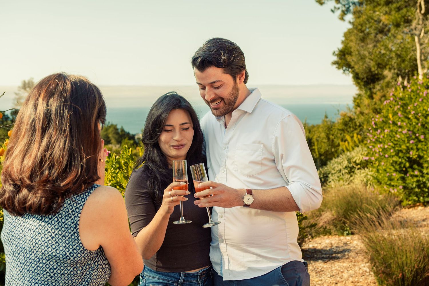Three friends toasting with champagne glasses outdoors at sunset with ocean views in the background.