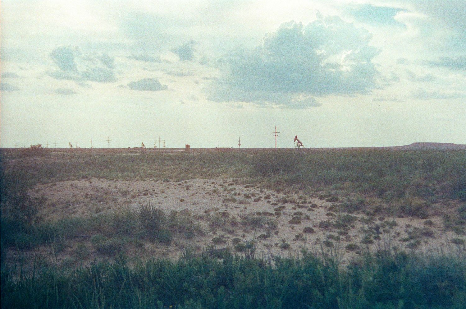 Vast flat prairie landscape with oil pump jacks on the horizon under a cloudy sky in a remote oil field.