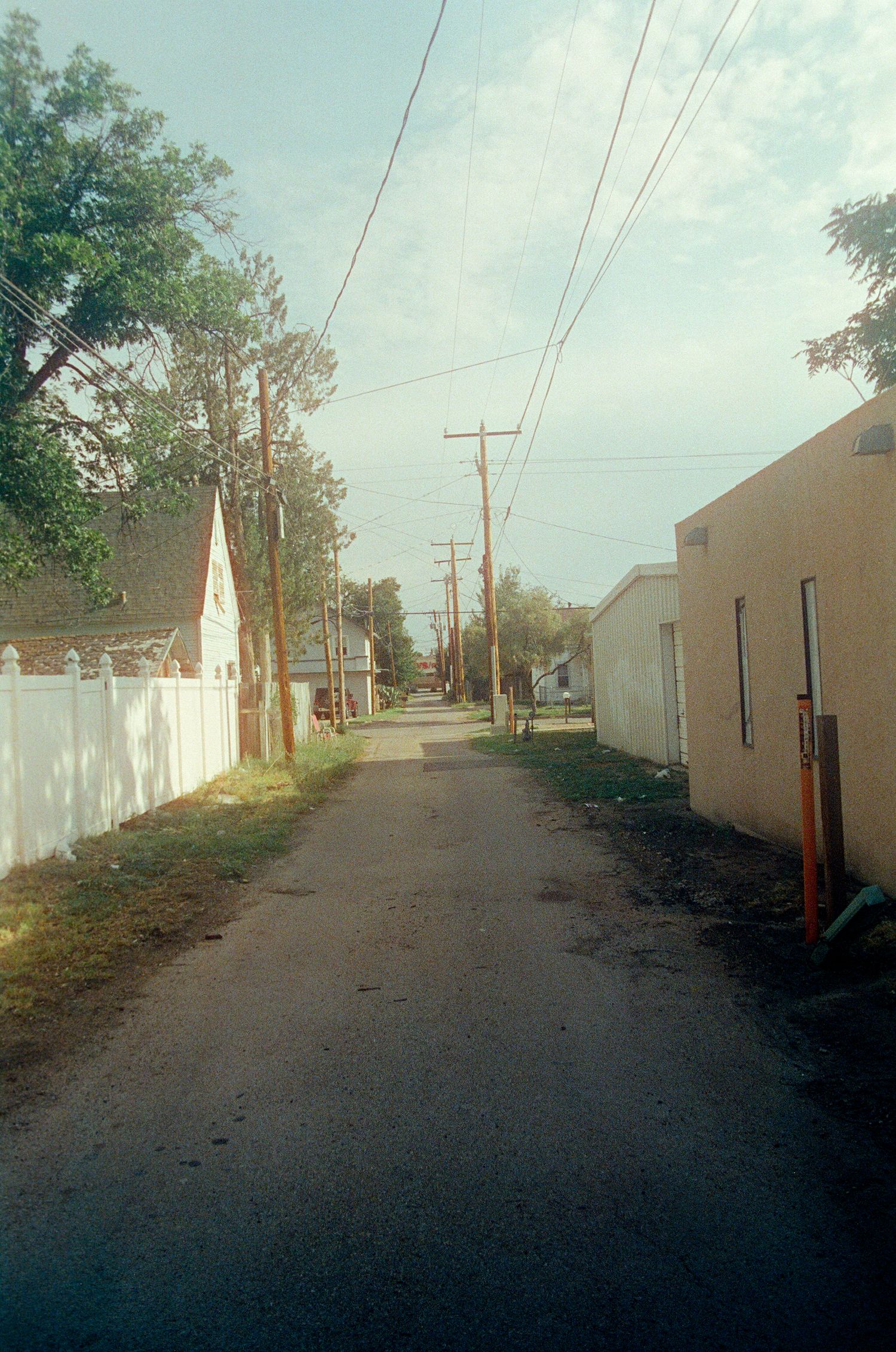 Narrow residential alley lined with power lines, white fence, and buildings under a partly cloudy sky.