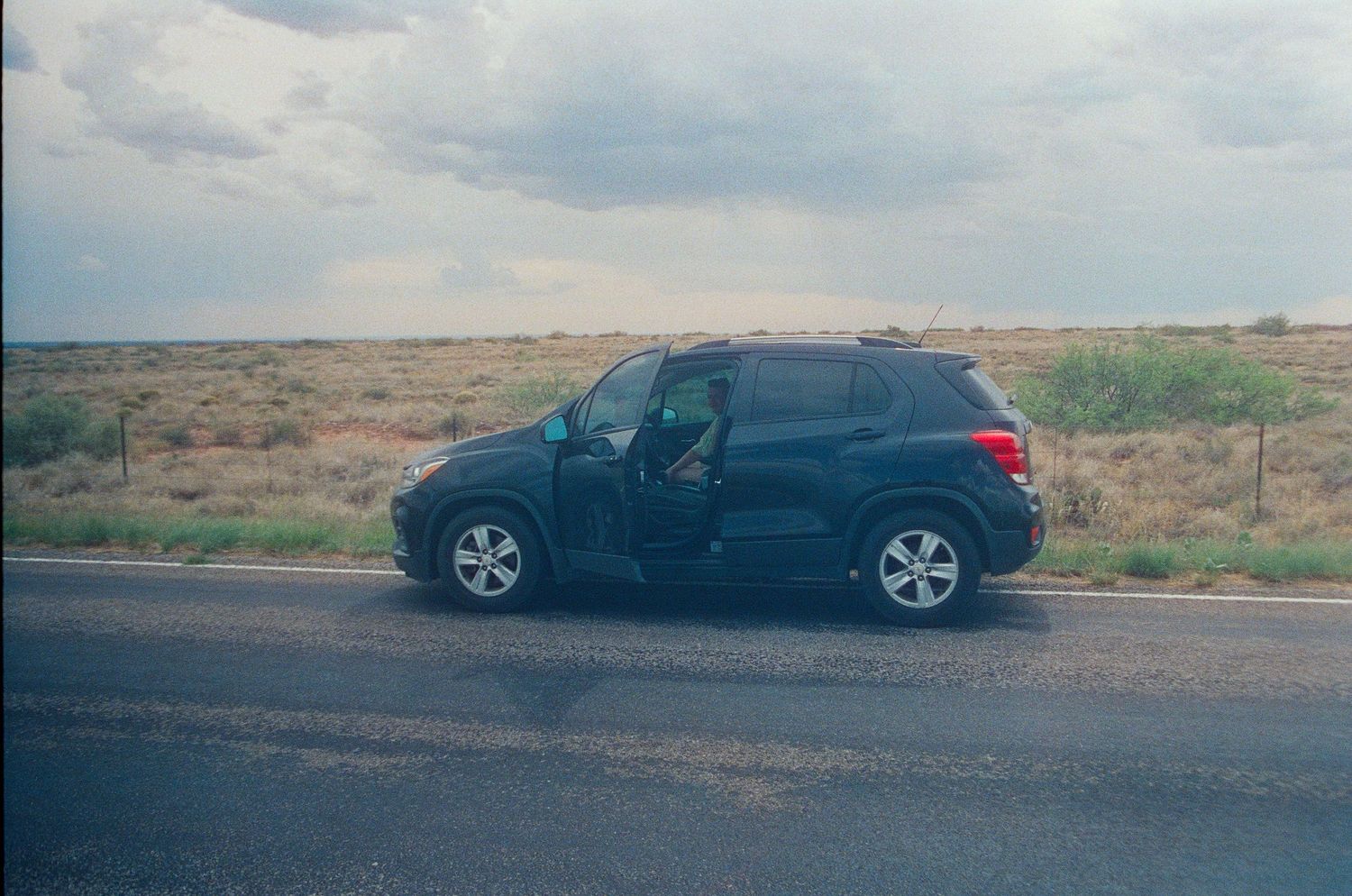 Black SUV parked on desert highway shoulder with open door, arid landscape and cloudy sky in background.