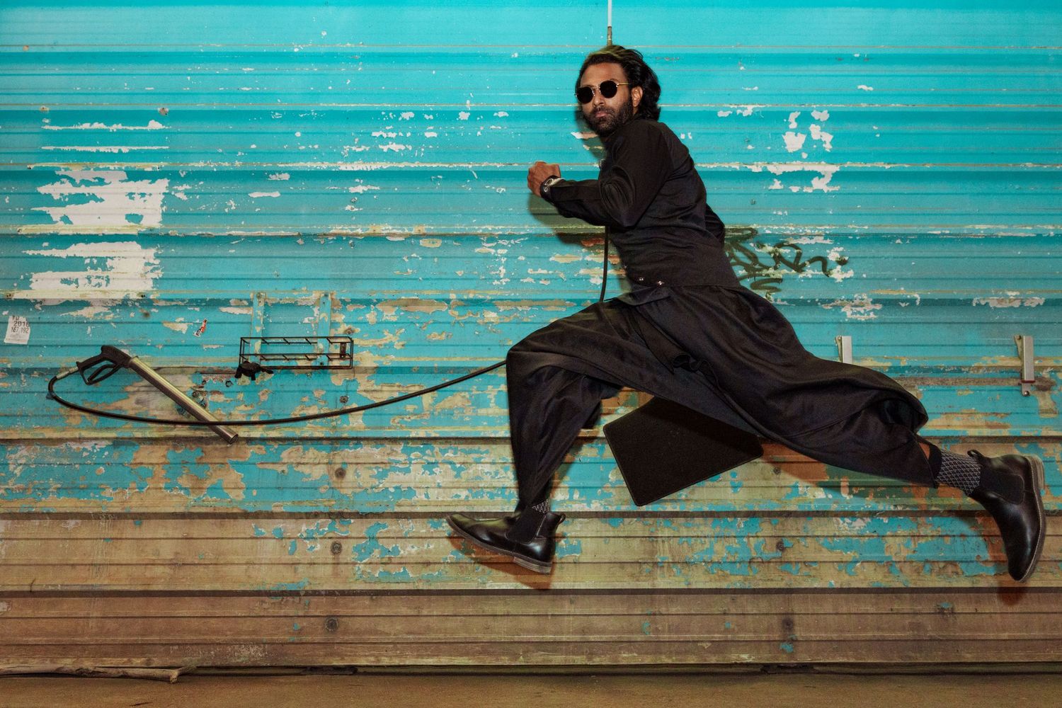 Dynamic action portrait of a man in flowing black attire leaping against a weathered turquoise wall.