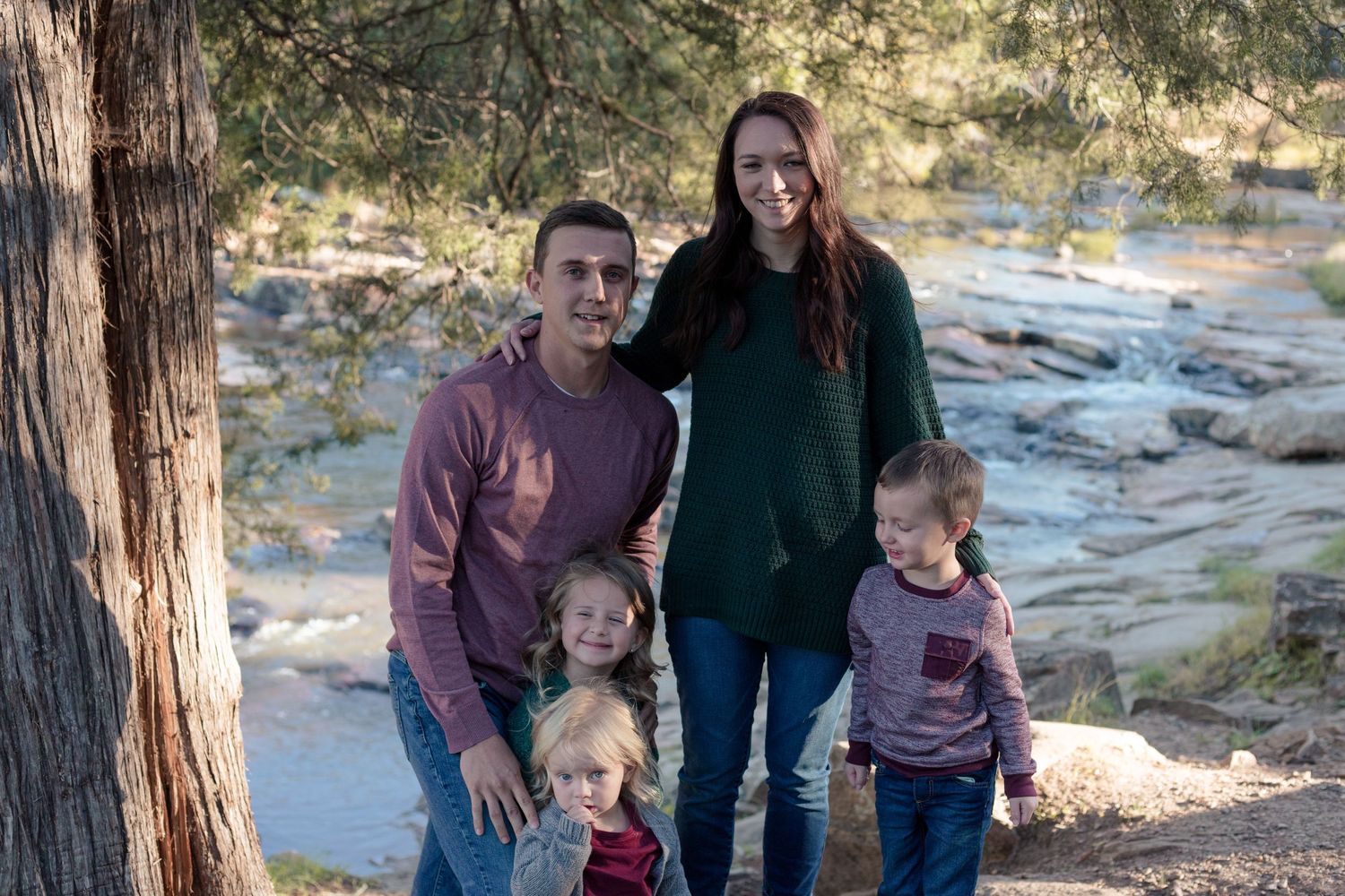 Family of five smiling together outdoors near a pine tree by a scenic river.