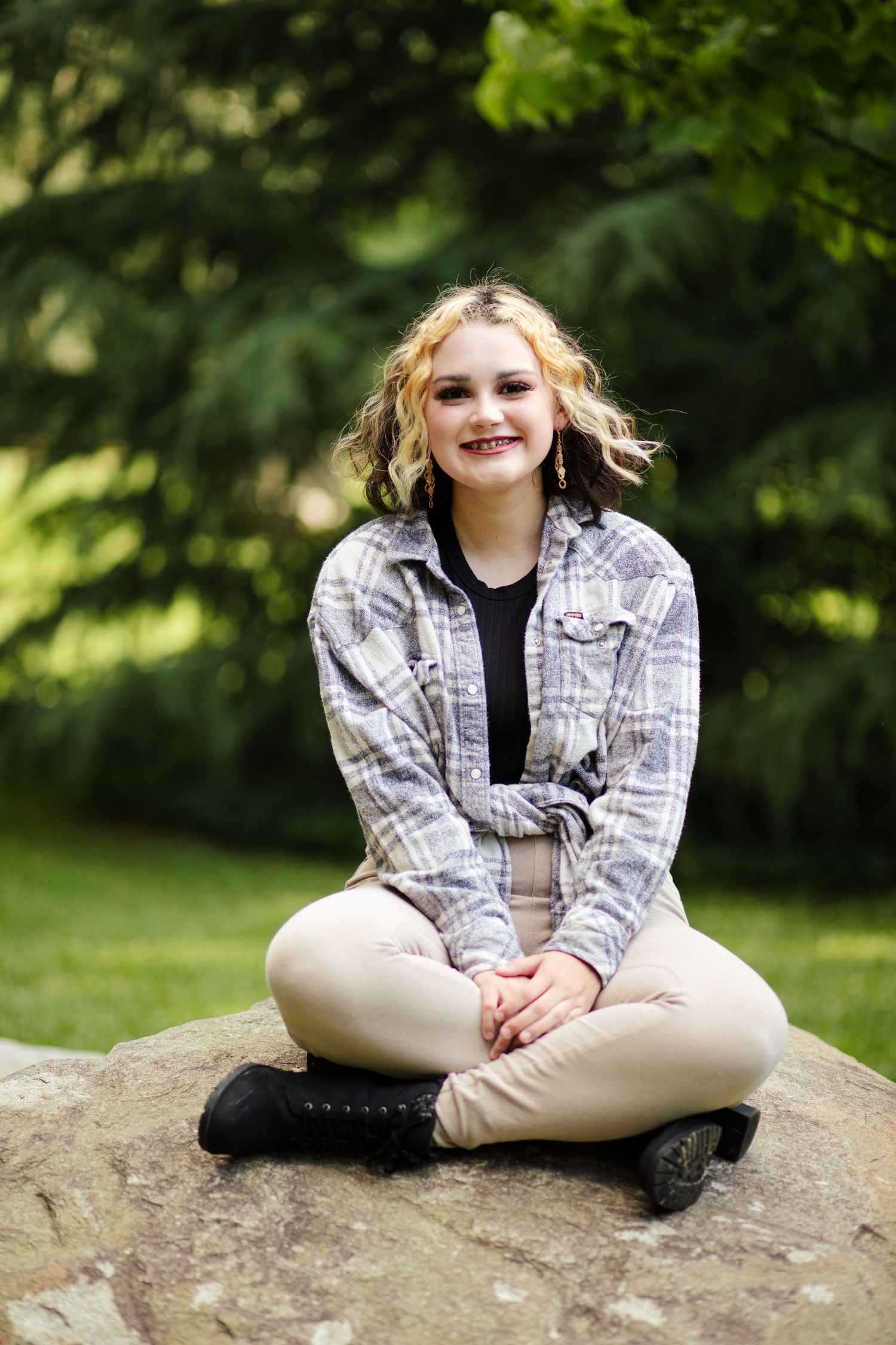 Smiling teenage girl in plaid shirt sitting cross-legged on rock in green park.