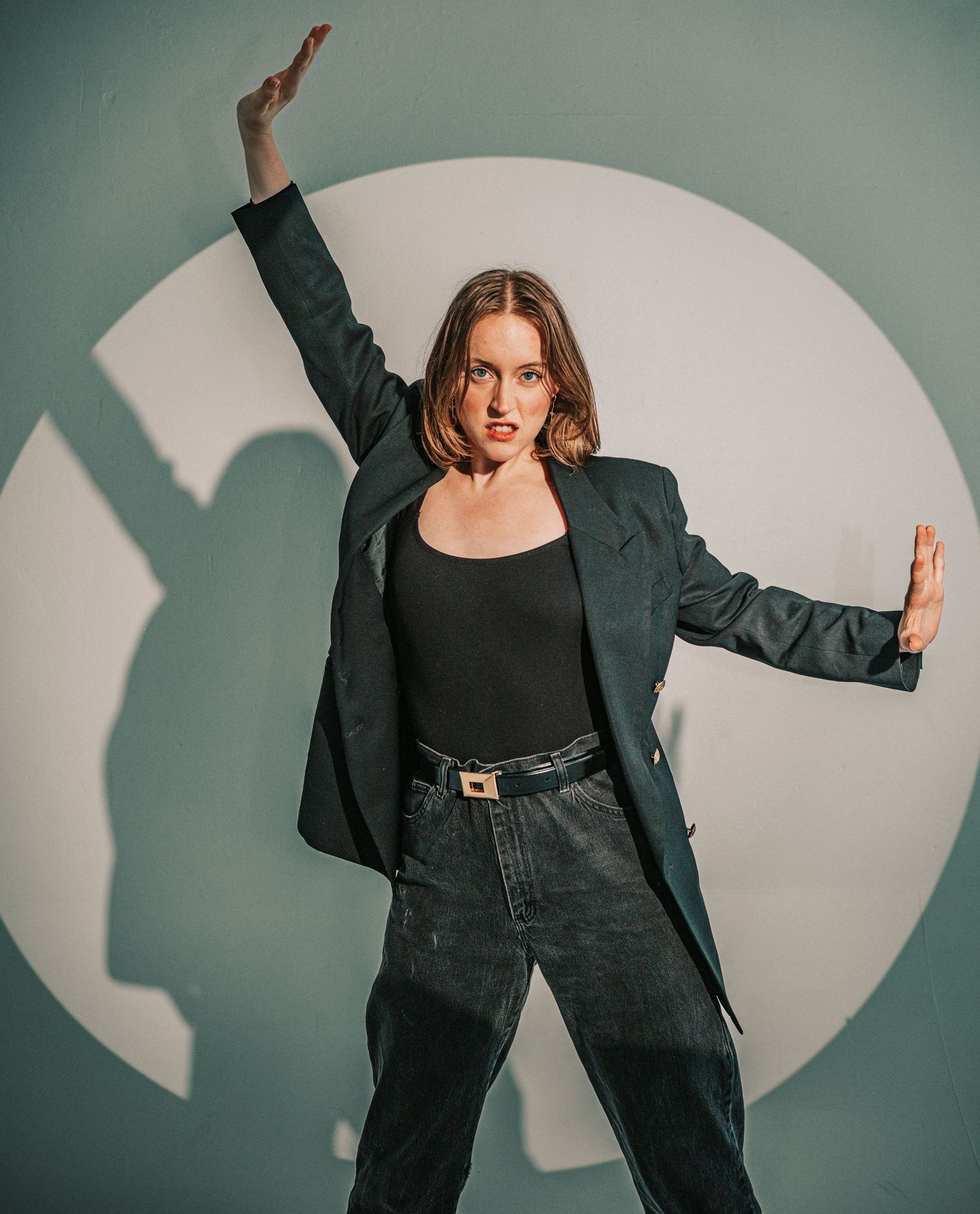 Confident woman in green blazer and jeans posing dramatically against circular white wall.