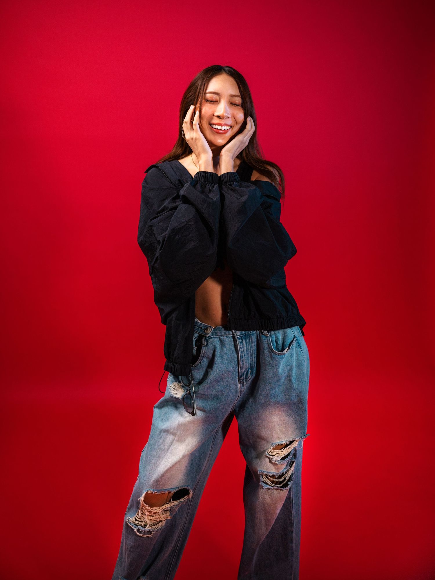 Cheerful Asian woman in black oversized top and ripped jeans smiles with hands on cheeks on red background.