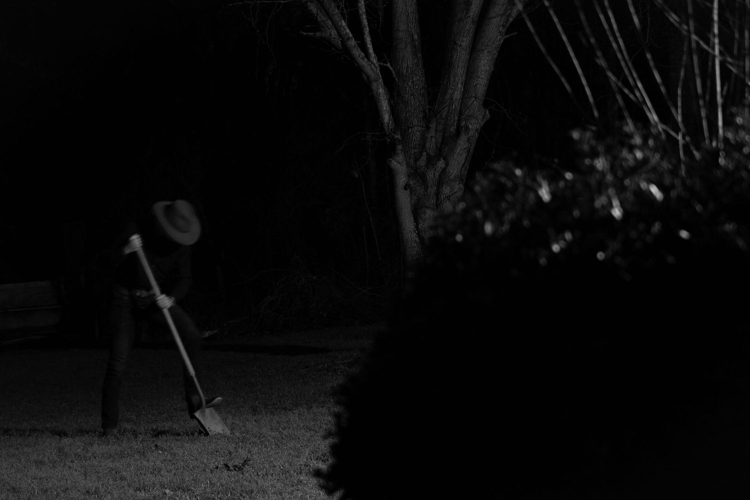 Black and white night photo of a person wearing a hat, raking leaves in a dark garden near trees.
