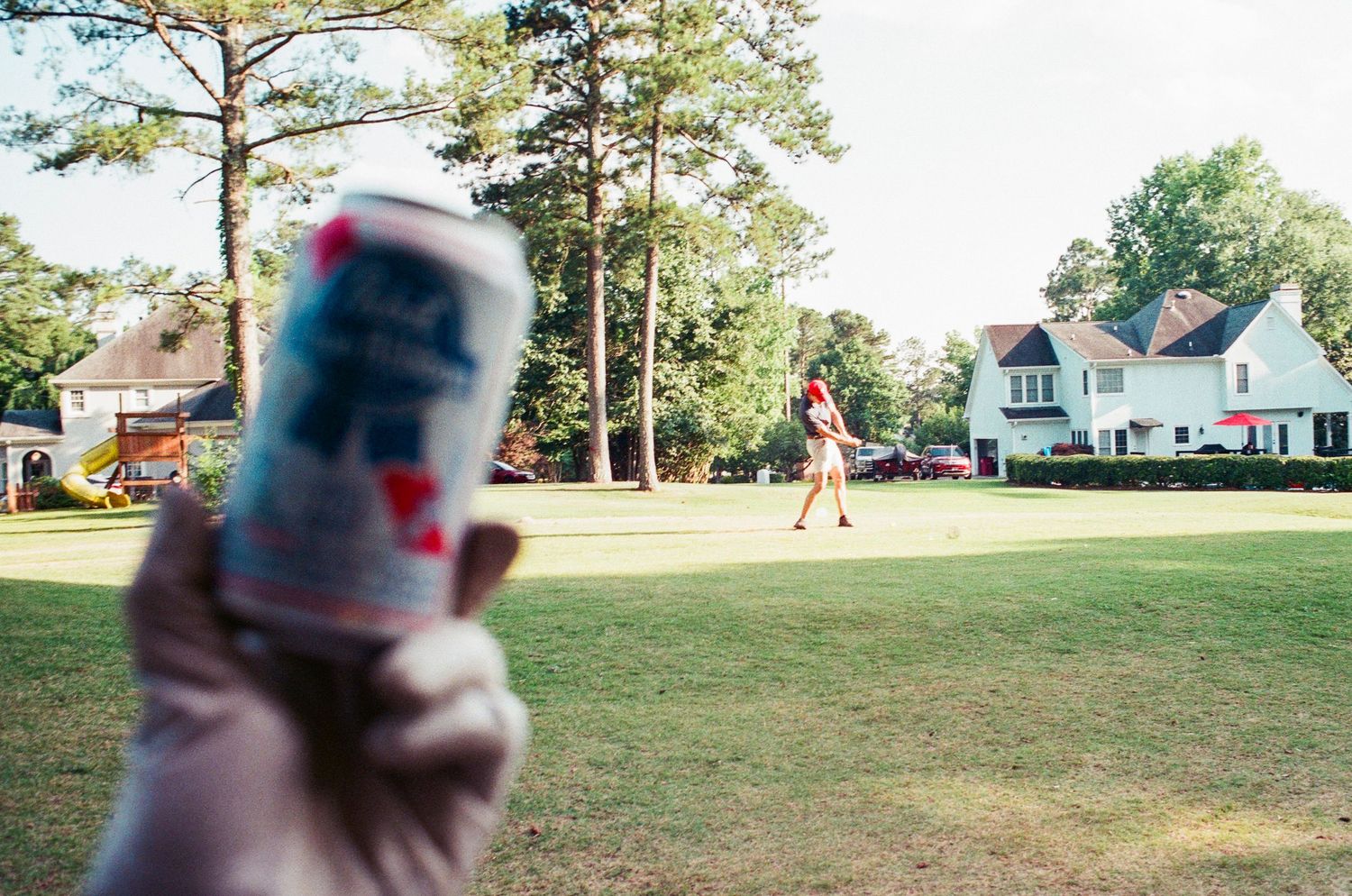 Person holding a Pabst Blue Ribbon beer can on a sunny golf course with a golfer and white house in background.
