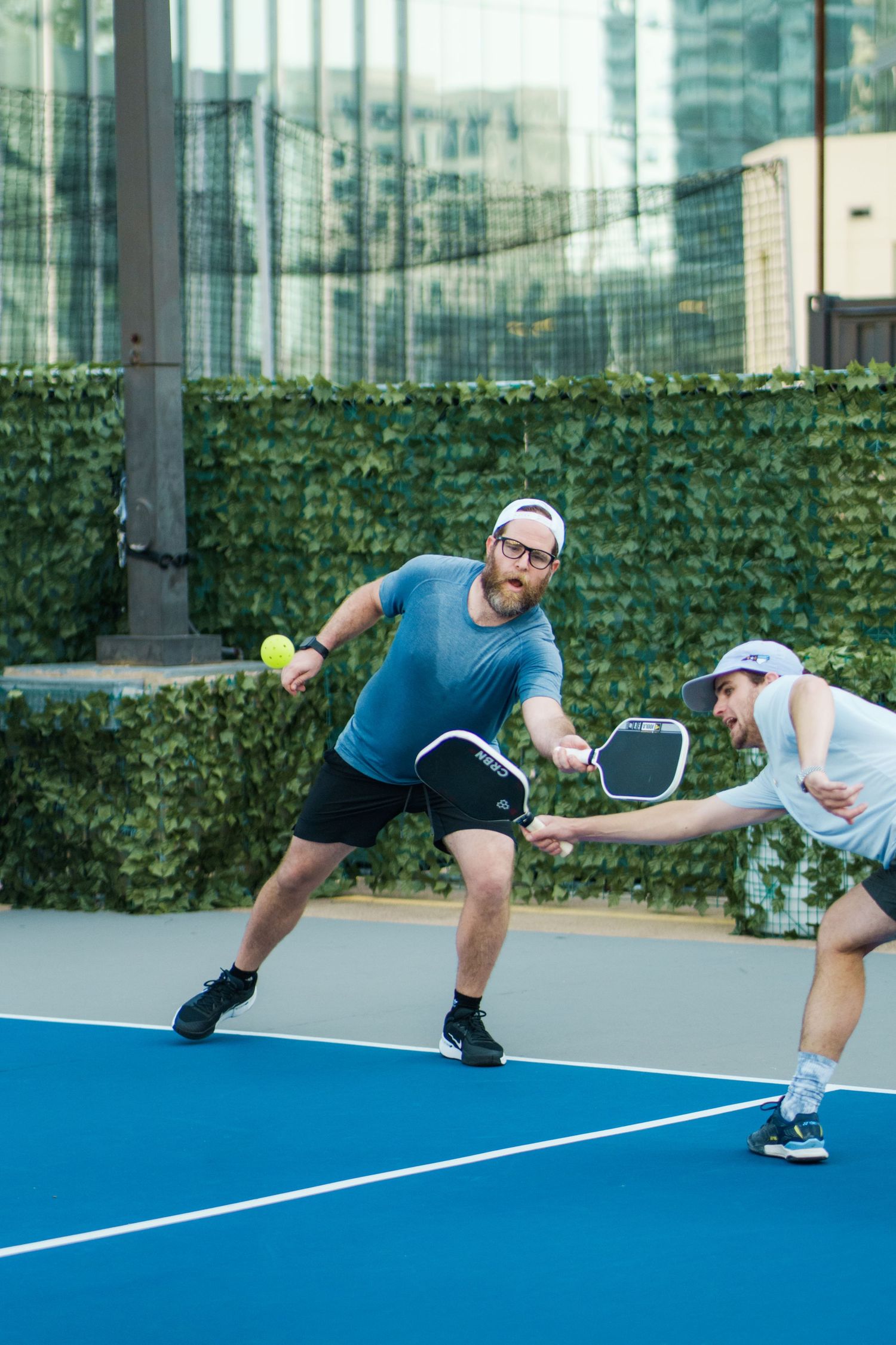 Two men playing pickleball on a blue outdoor court, surrounded by green ivy walls and urban buildings.