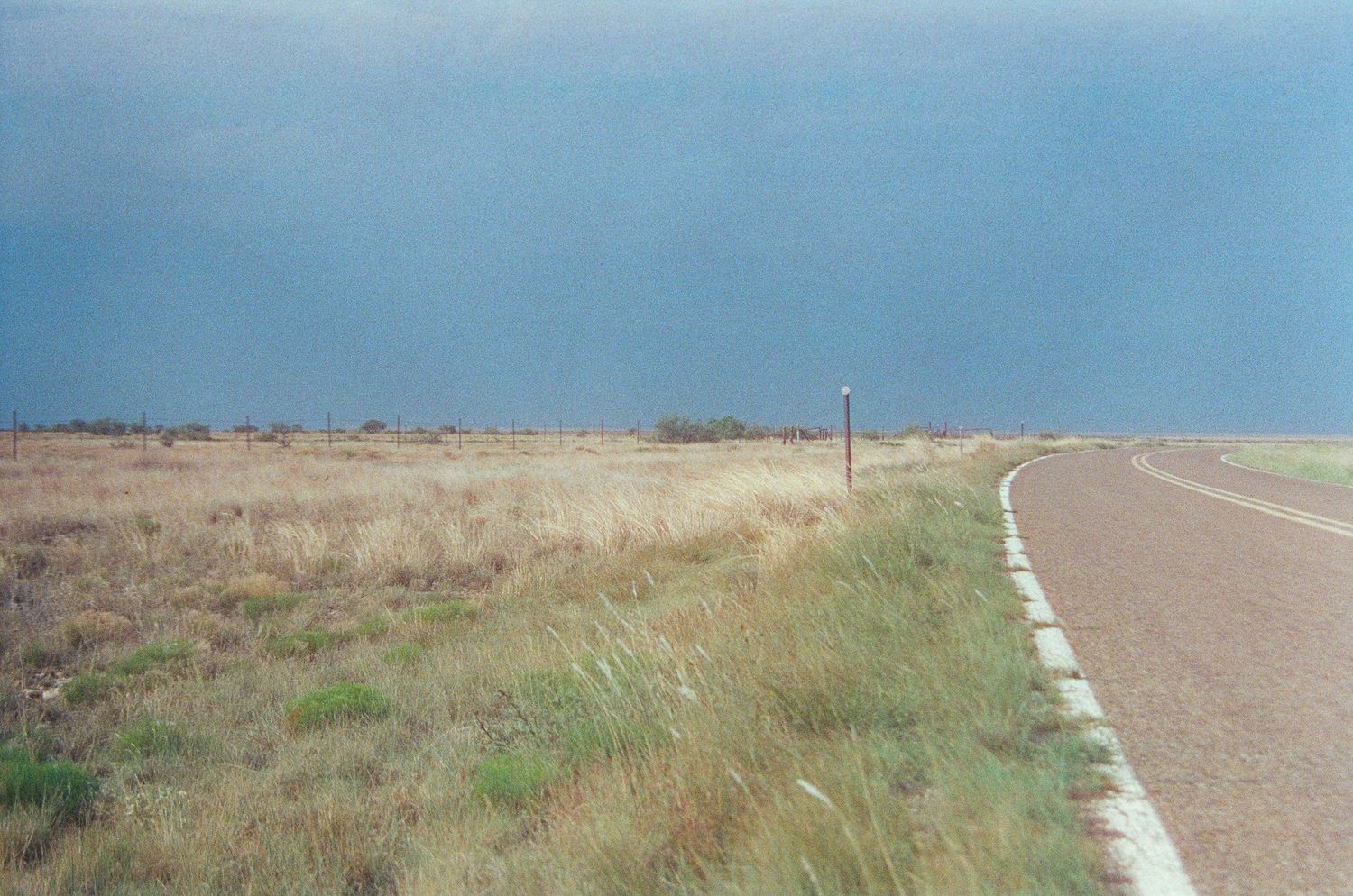 A desolate rural highway stretches through flat grassy plains under a stormy blue-gray sky in the American Midwest.