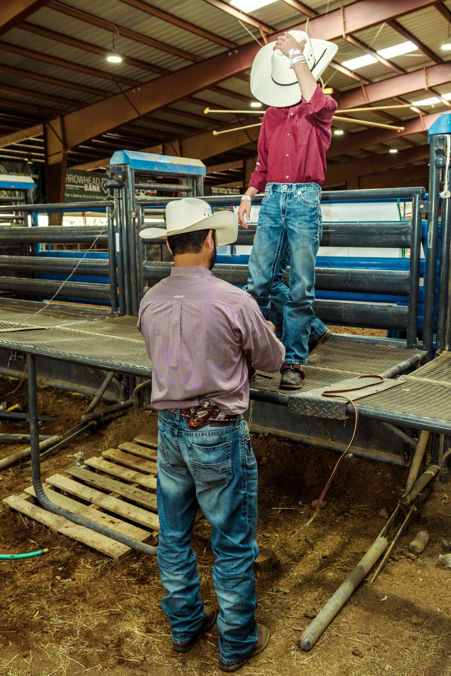 Two cowboys in western shirts, jeans, and white hats prepare near rodeo chutes inside an indoor arena.