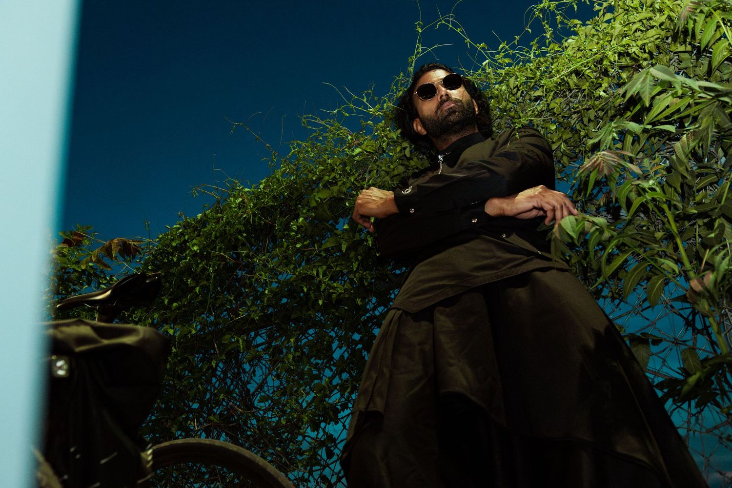 Editorial portrait of a man in black attire posing beside a bicycle amid lush green foliage and blue sky.