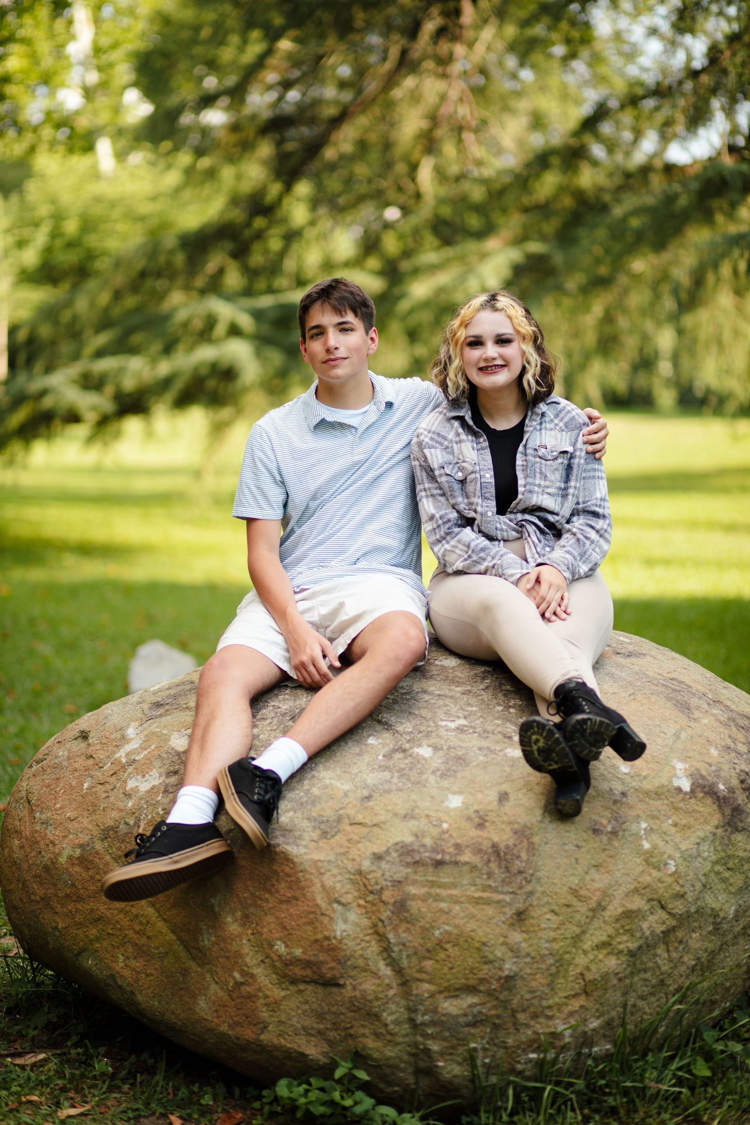 Teen boy and curly-haired girl posing together on large rock in green park.