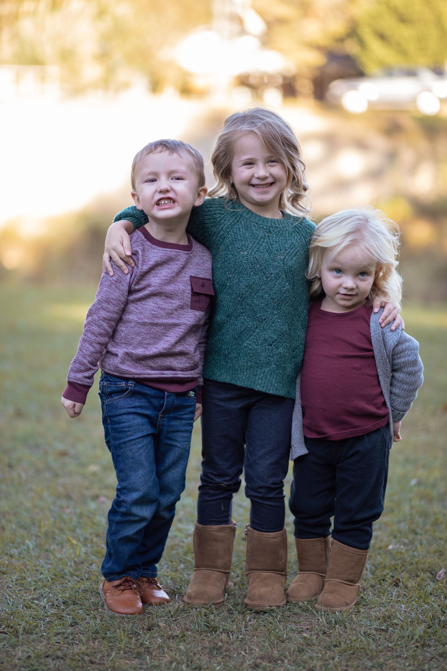 Three young siblings hugging and smiling together in a sunny outdoor park setting.