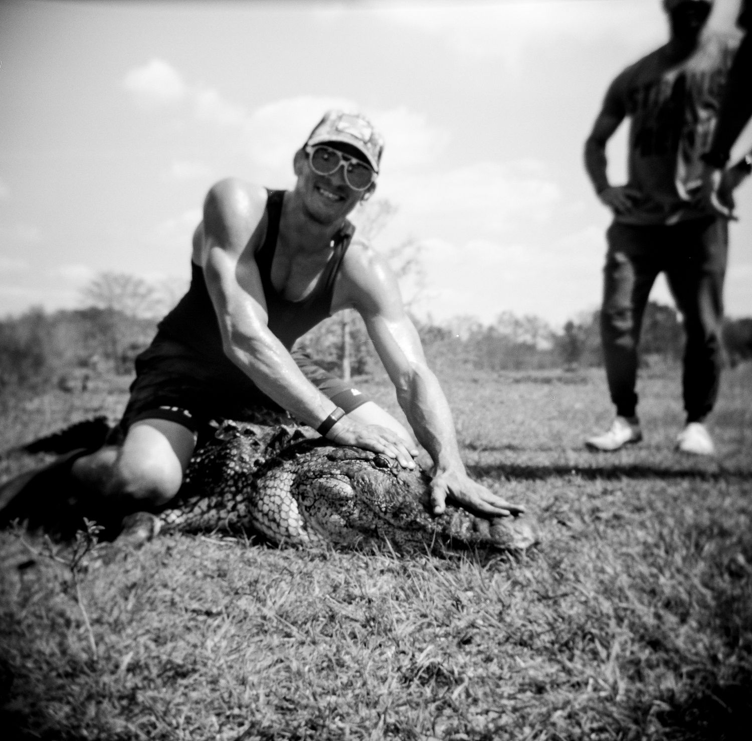 Muscular man in cap and sunglasses wrestles an alligator on grass in black and white photo.