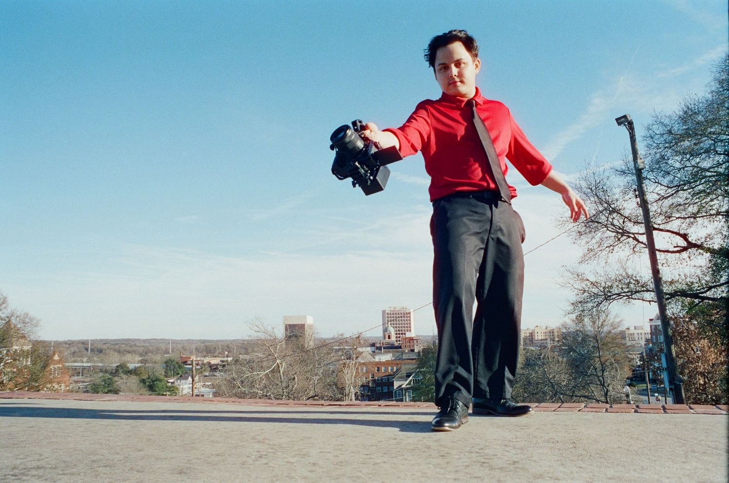 Person in red shirt holding camera low angle shot with city skyline behind.