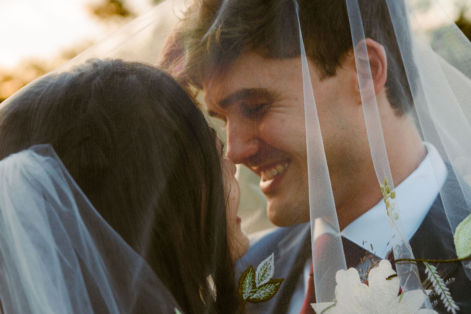 A smiling groom and bride share an intimate moment under a white wedding veil outdoors.