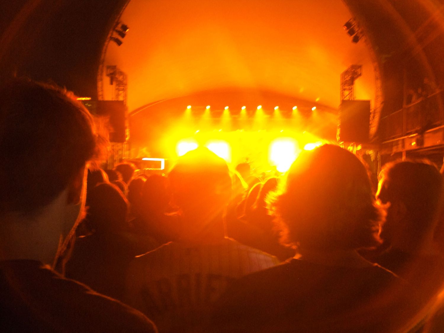 Crowd at an outdoor concert bathed in warm orange stage lights at night.