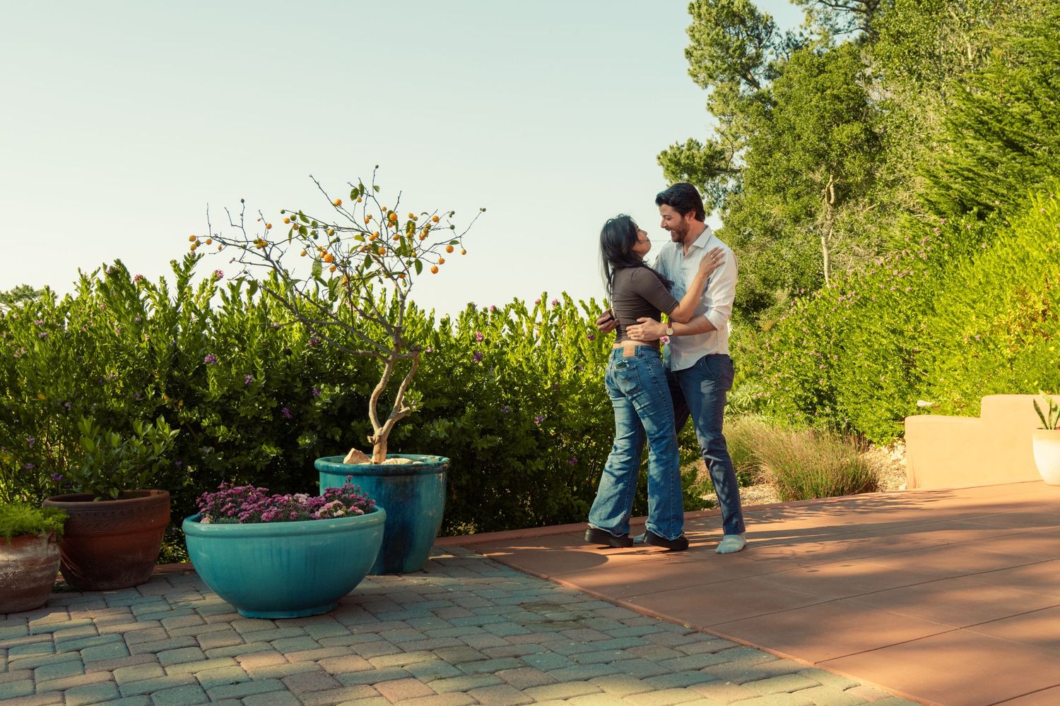 Couple dancing together on a brick patio surrounded by lush green plants and potted trees in a garden setting.