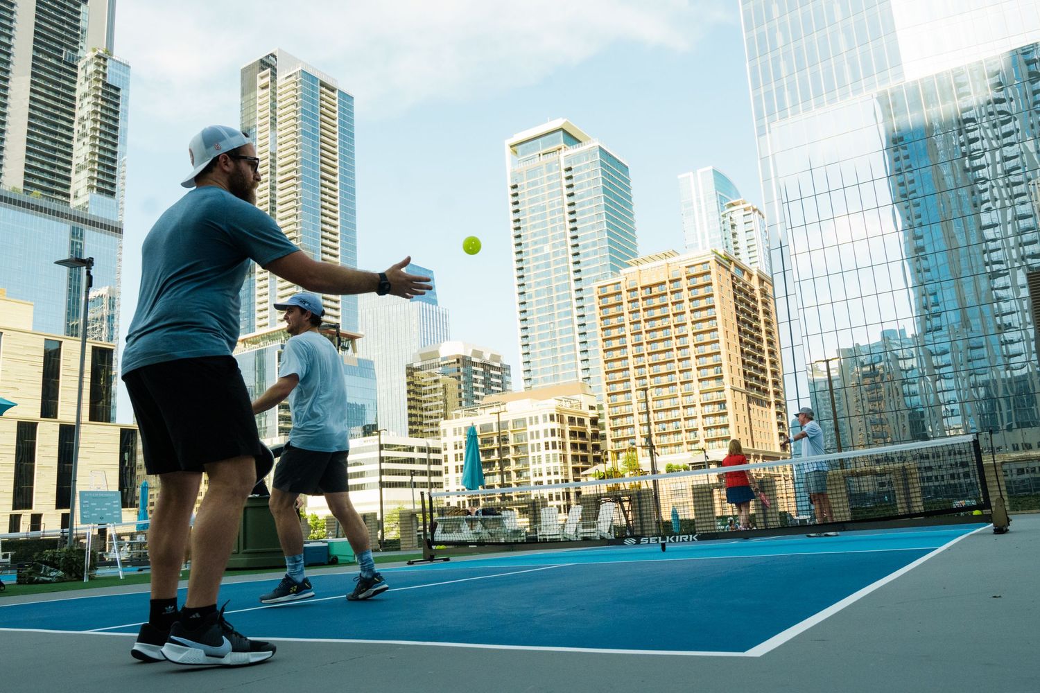 Players enjoy a pickleball game on a rooftop court surrounded by modern city skyscrapers on a sunny day.