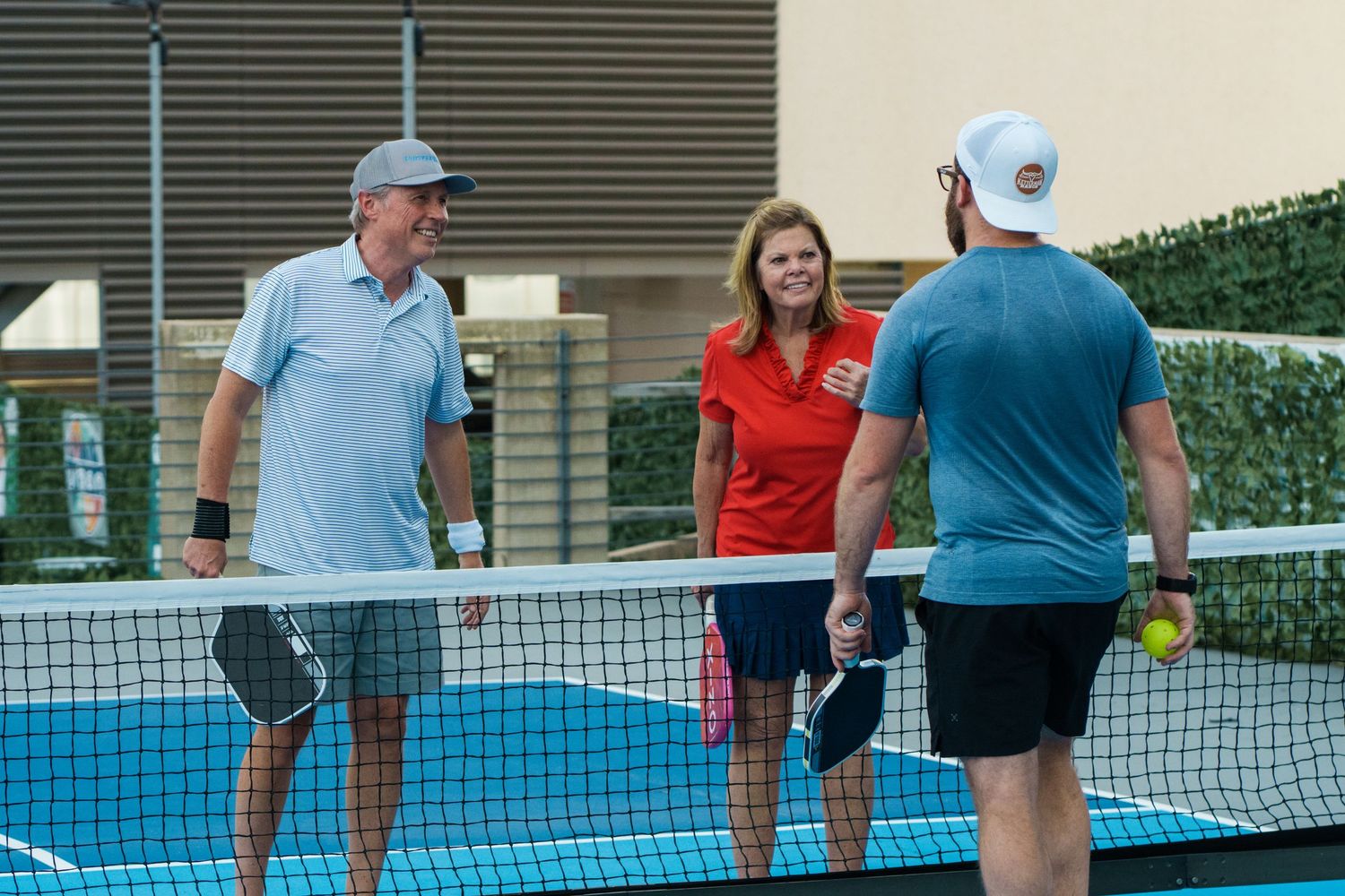 Three adults playing pickleball on an outdoor court, two men and a woman near the net holding paddles.