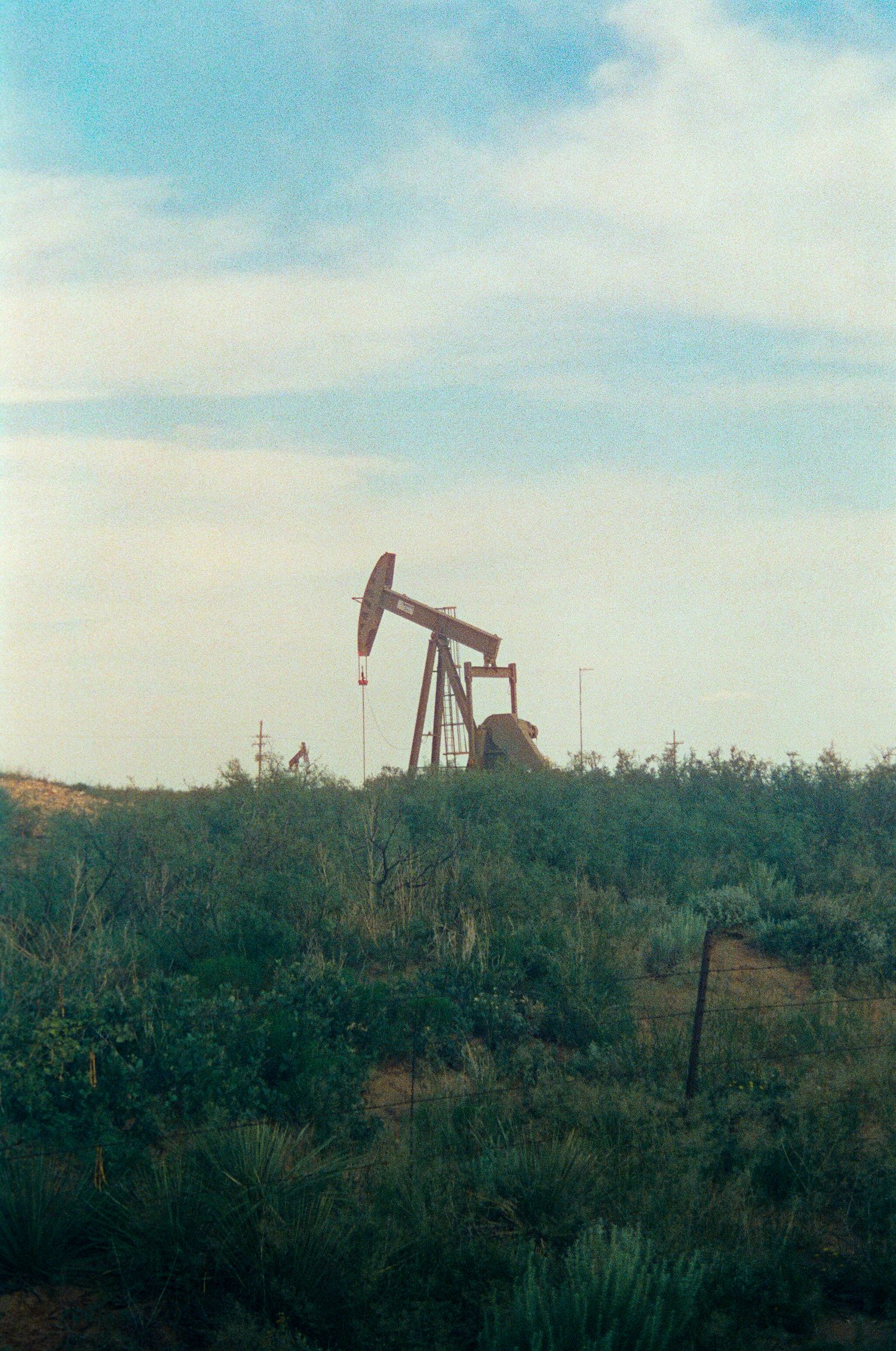 Oil pump jack operating in a vast green field under a partly cloudy blue sky in a rural oil field.