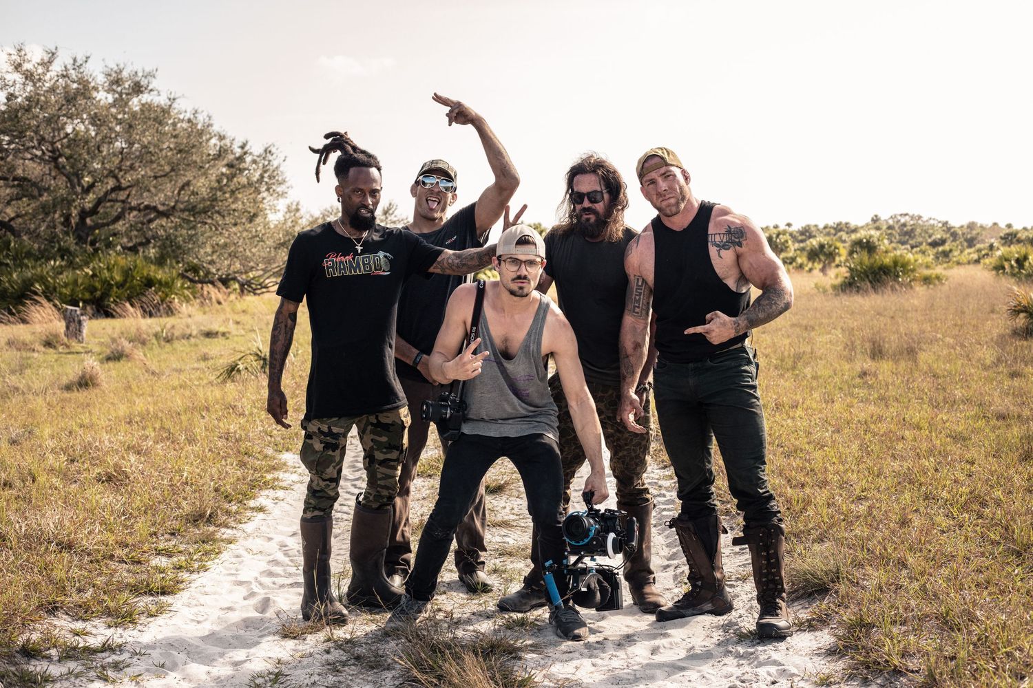 Five diverse men posing together on a sandy trail in a sunny, grassy outdoor setting, one holding camera equipment.