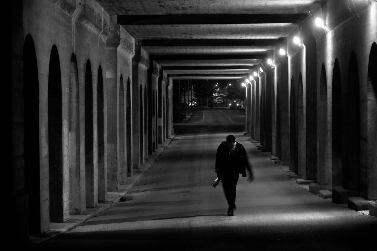 A lone man walks through a dimly lit arched corridor at night in black and white photography.