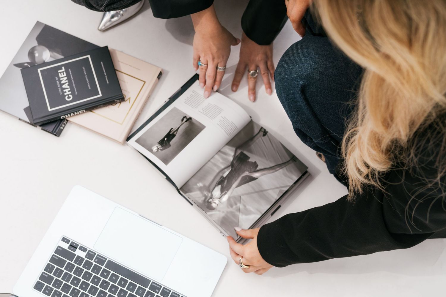 Blonde woman reviews a photography magazine beside a laptop and notebooks on a white desk.