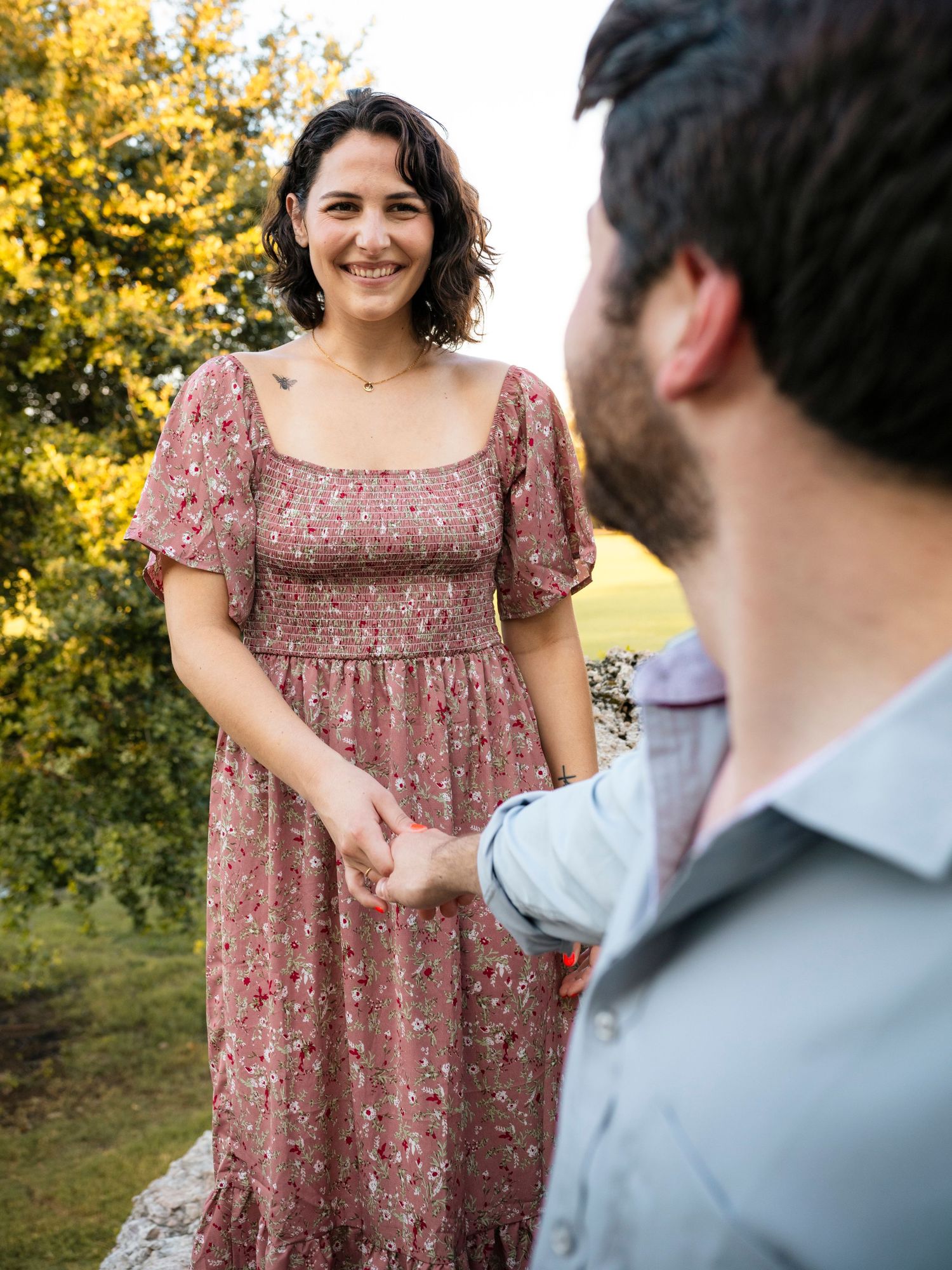 Smiling woman in floral dress holds hands with bearded man during outdoor engagement session.