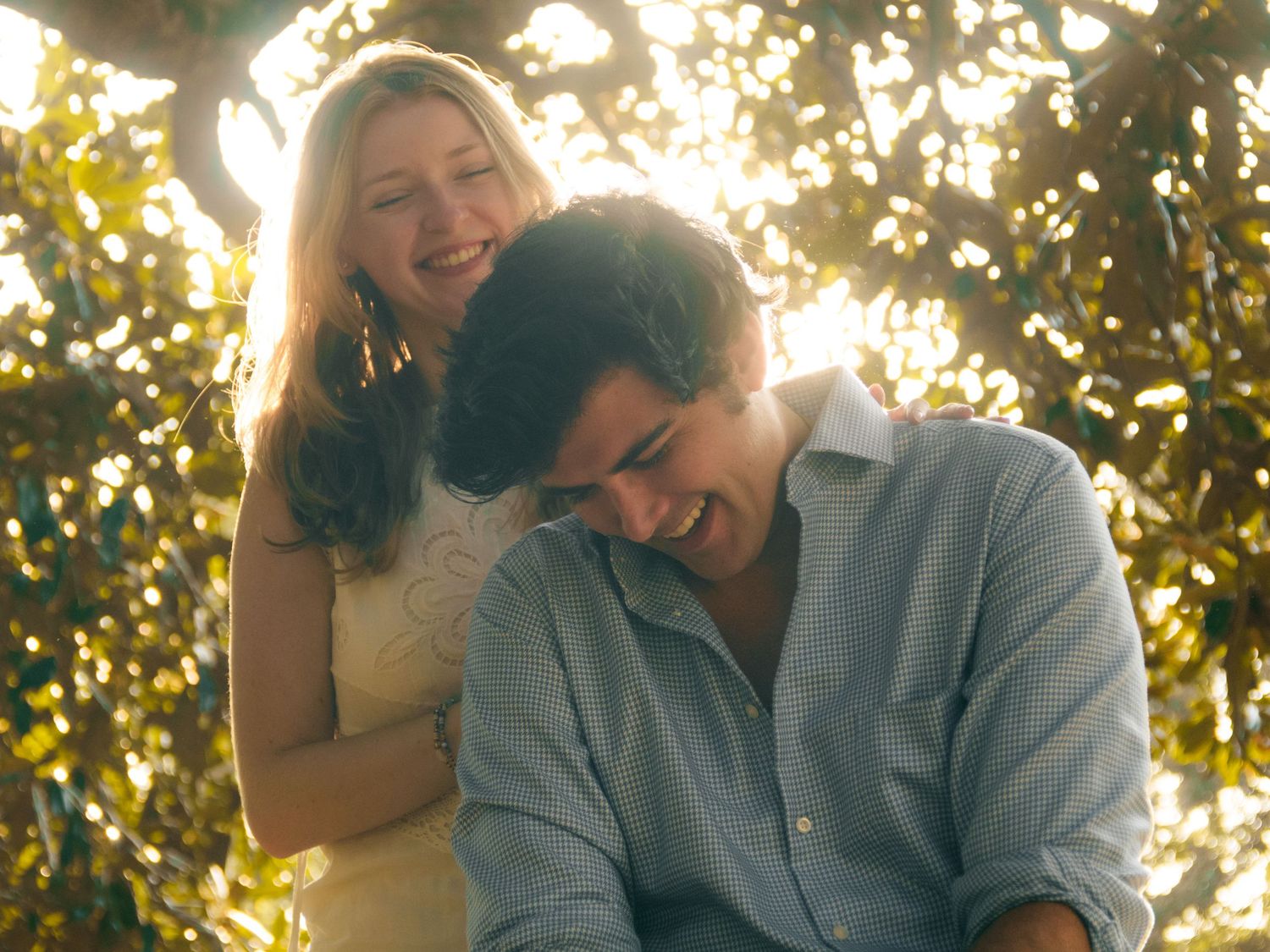 Joyful couple laughing together outdoors with golden sunlight filtering through green foliage.