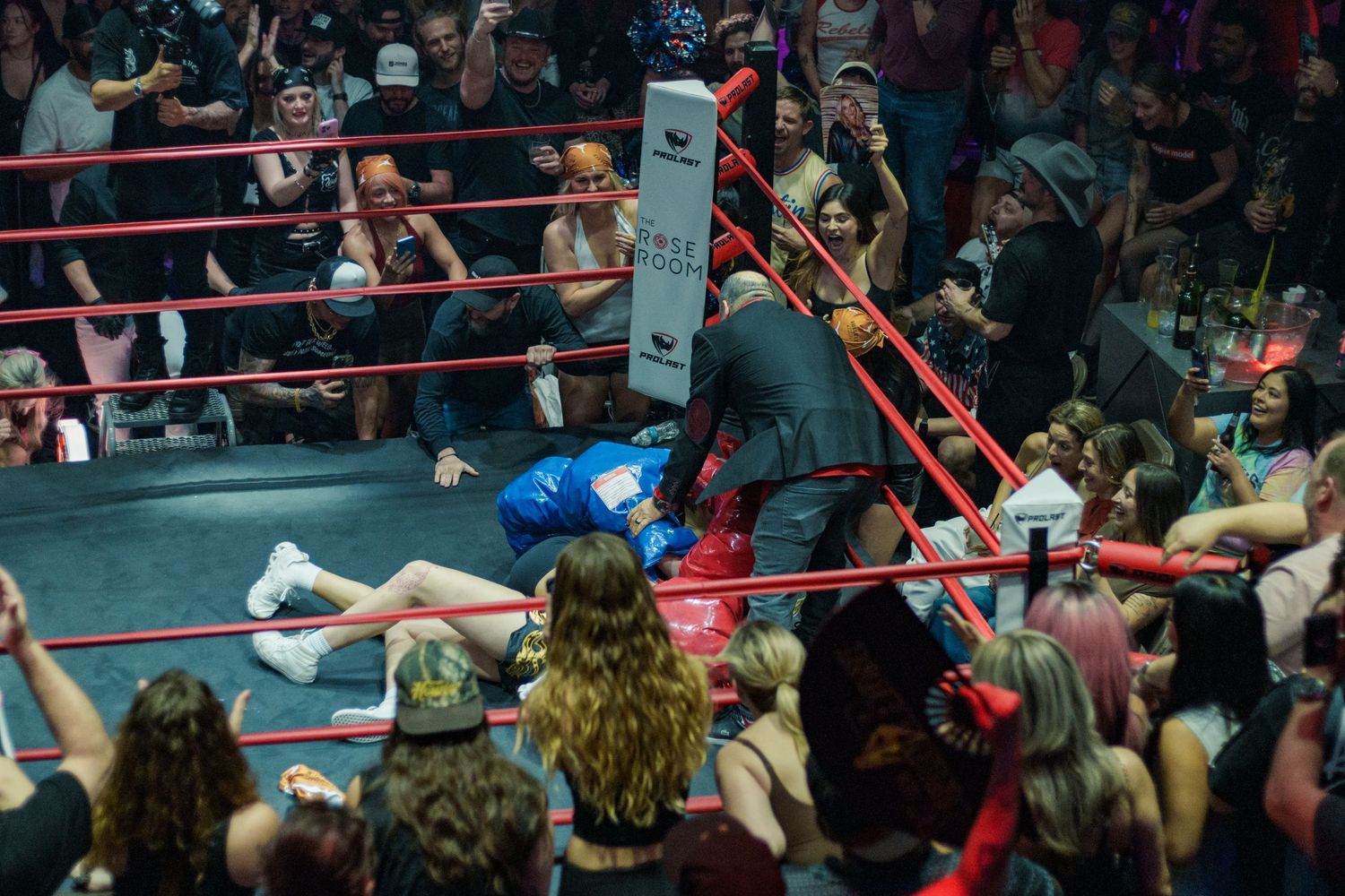 A female boxer in blue gloves falls near the ropes as a referee intervenes in a packed boxing event.