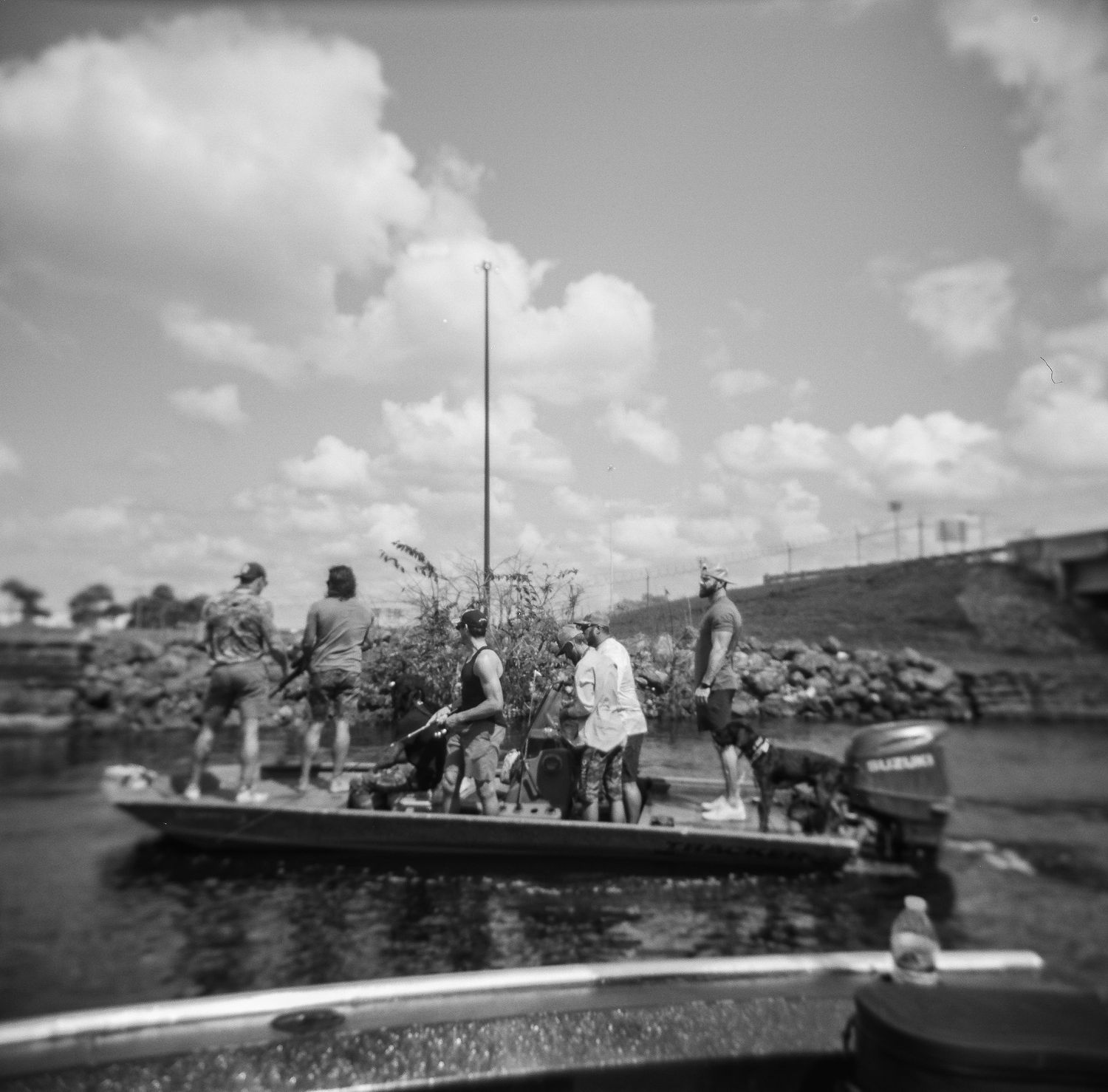 A film crew on a boat near a rocky shoreline with an outboard motor, black and white photo.