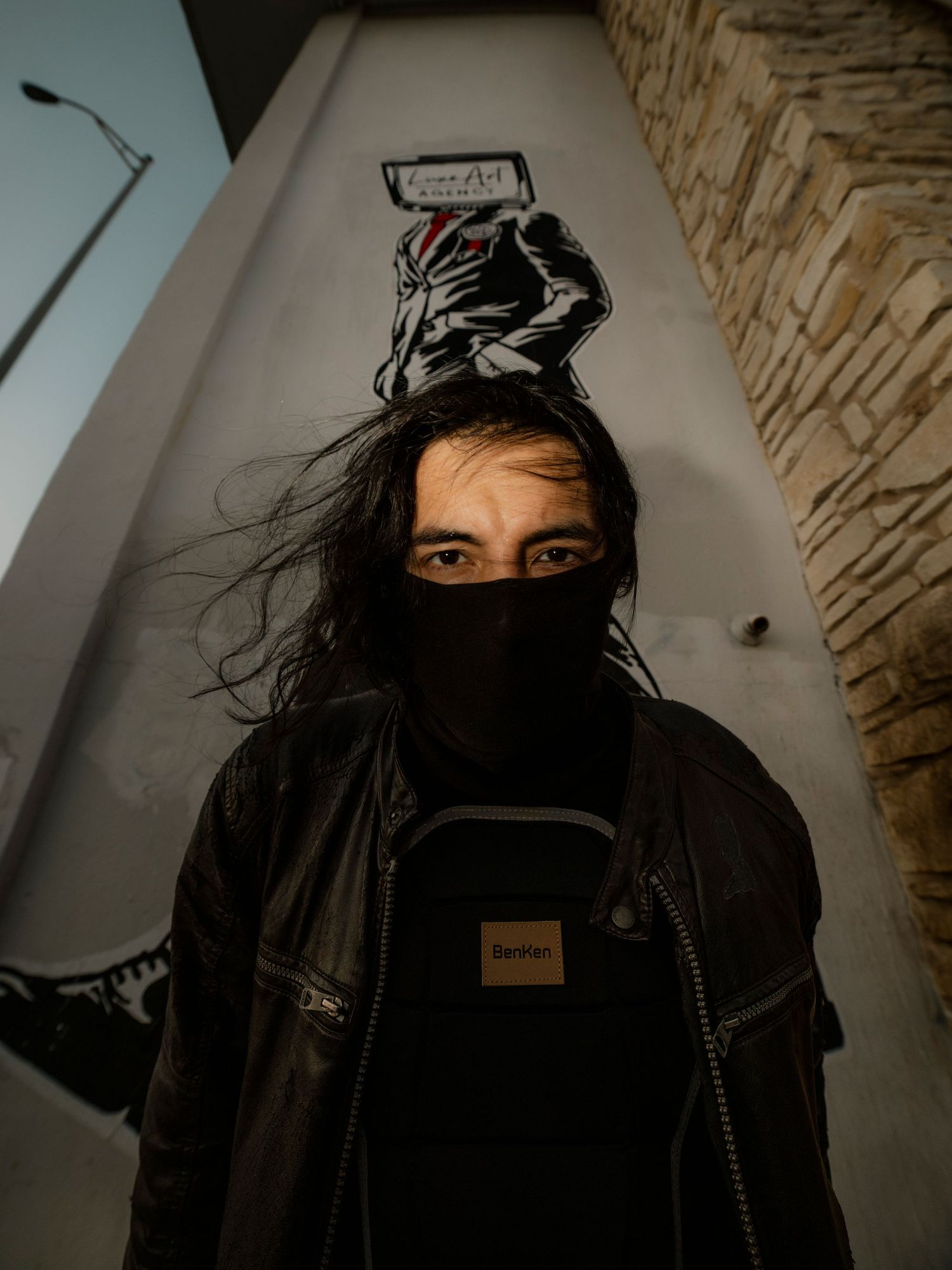 Long-haired person in black mask and leather jacket standing beneath street art mural.