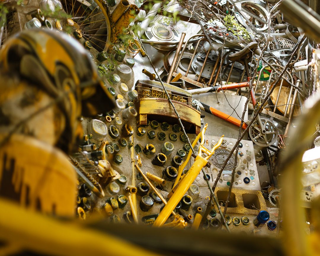 Cluttered junkyard pile of scrap metal, yellow machinery parts, wires, and glass bottles in an industrial salvage yard.
