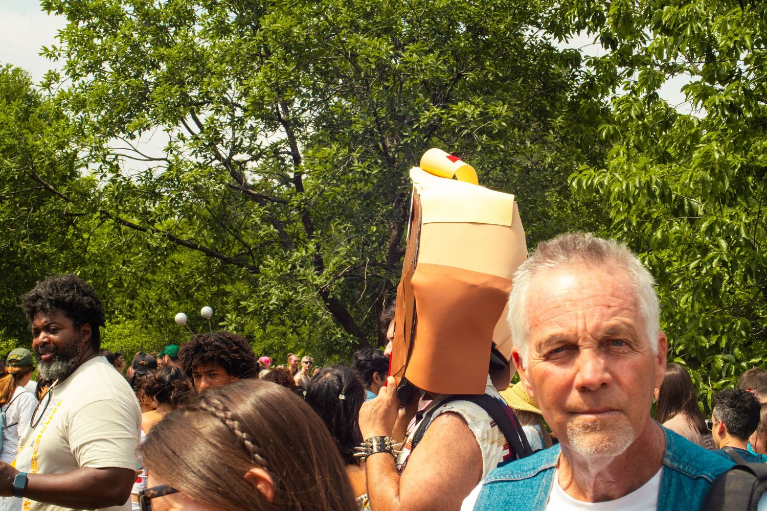 Crowd at outdoor festival with man in foreground and woman holding large drink cup above her head, green trees behind.