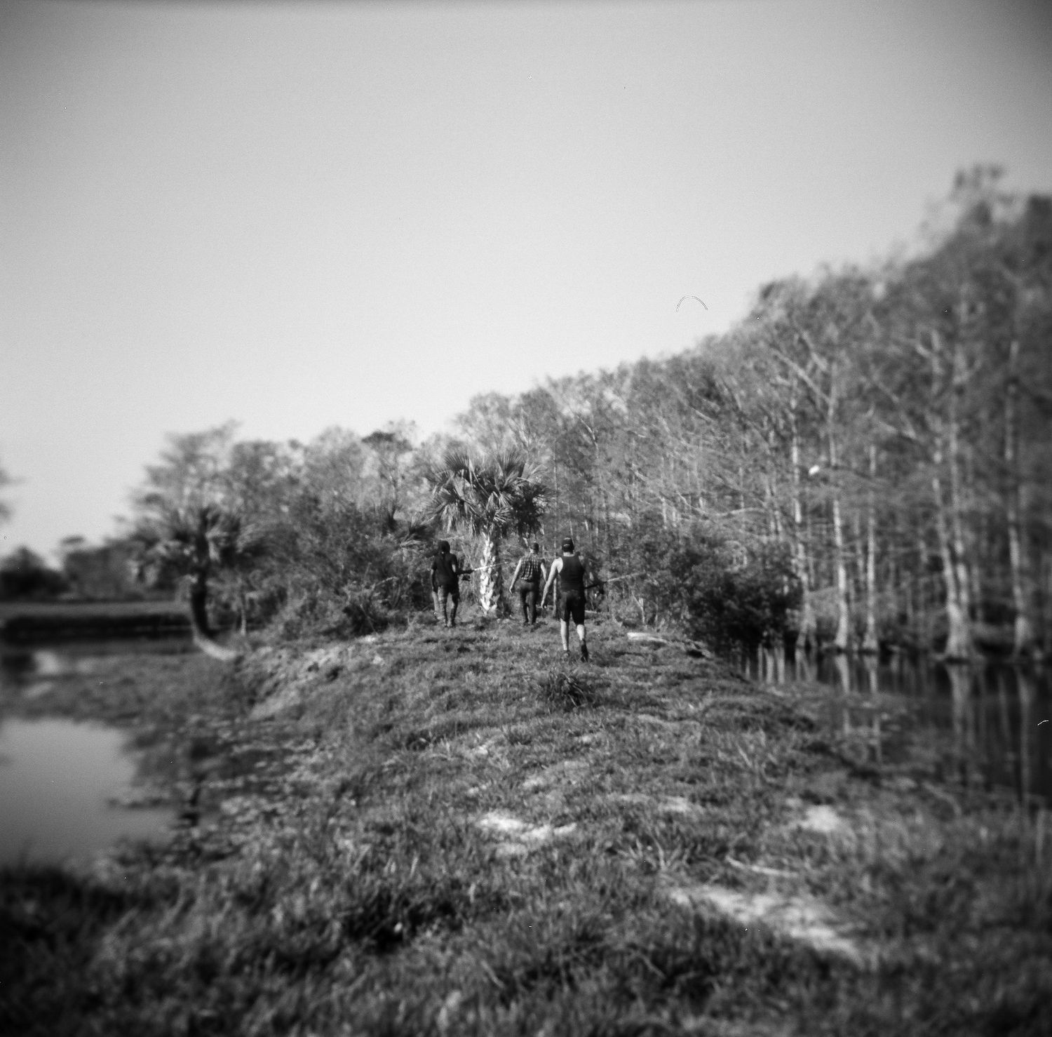 Black and white photo of people walking along a grassy path beside a river, surrounded by tall trees.