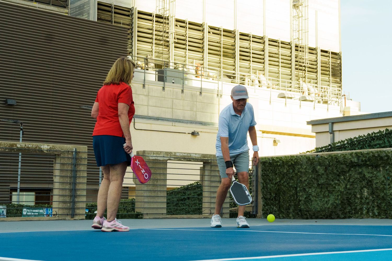 Two players practice pickleball on a blue outdoor court, with a woman in red and a man in a hat mid-swing.